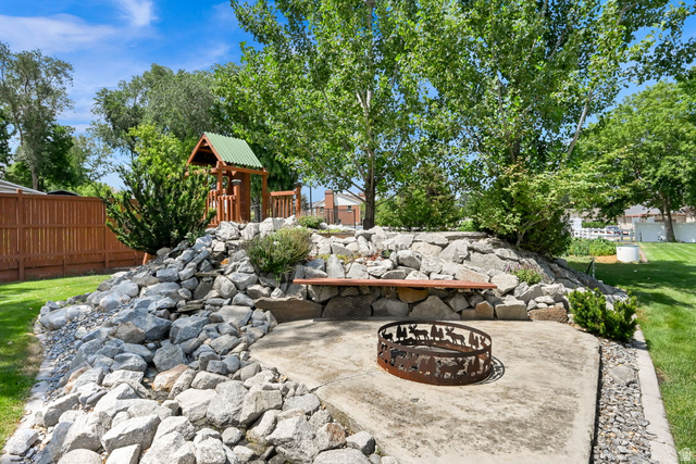 13658 South Fort Street Draper, UT 84020 - Photo 64 of 87 Outdoor entertainment area featuring a concrete patio with a decorative fire pit, a built-in wooden bench, a rock retaining wall, and mature trees