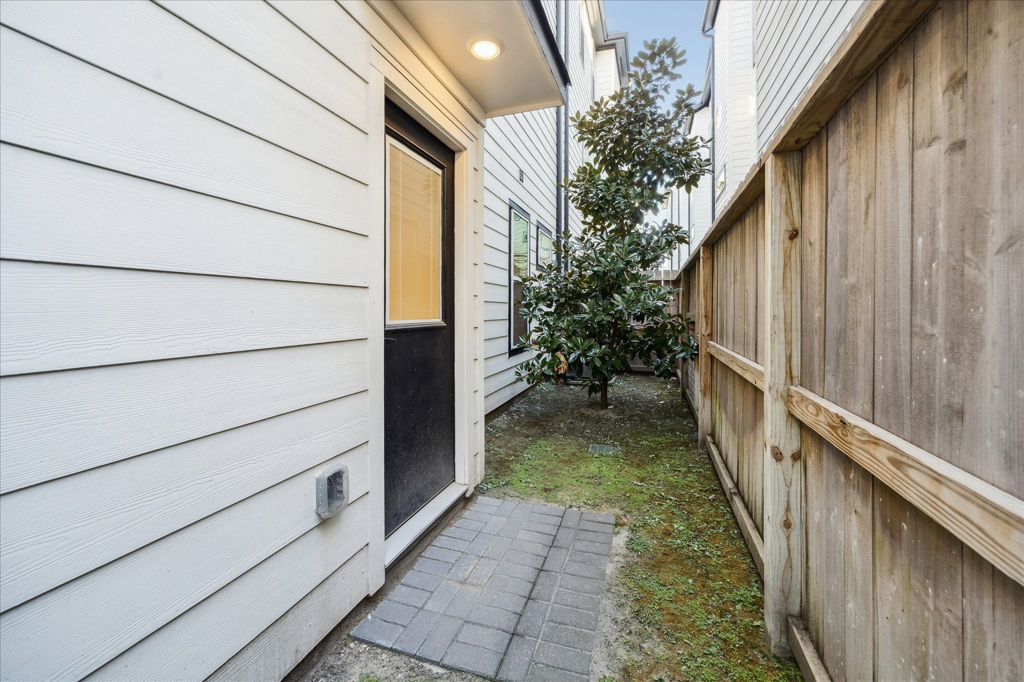 1603 Patterson Street, Unit E Houston, TX 77007 - Photo 31 of 32 This photo shows a narrow side yard with a paved walkway leading to an exterior door. The area is bordered by a wooden fence on one side and the house siding on the other, with a small tree adding greenery.