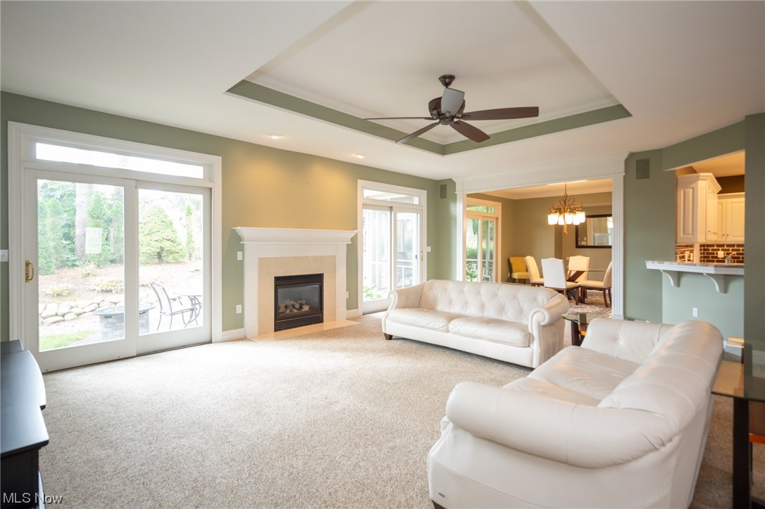 Living room featuring a raised ceiling, light colored carpet, a healthy amount of sunlight, and ceiling fan with notable chandelier