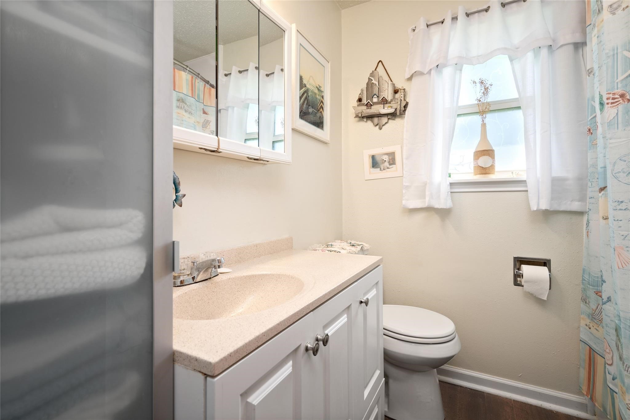 303 North 9th Street Beasley, TX 77417 - Photo 39 of 41 This cozy bathroom features a light-colored vanity with a sink, a toilet, and a window with sheer curtains allowing natural light. It includes a wall-mounted cabinet for storage and decorative coastal-themed elements.
