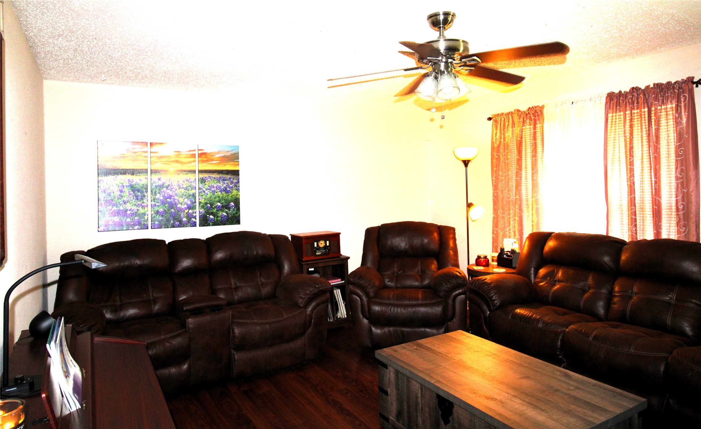 303 North 9th Street Beasley, TX 77417 - Photo 4 of 41 This cozy living room features dark leather sofas, a wooden coffee table, and a ceiling fan. Soft natural light filters through the curtains, complementing the warm, inviting atmosphere. A vibrant landscape painting adds a touch of color to the space.