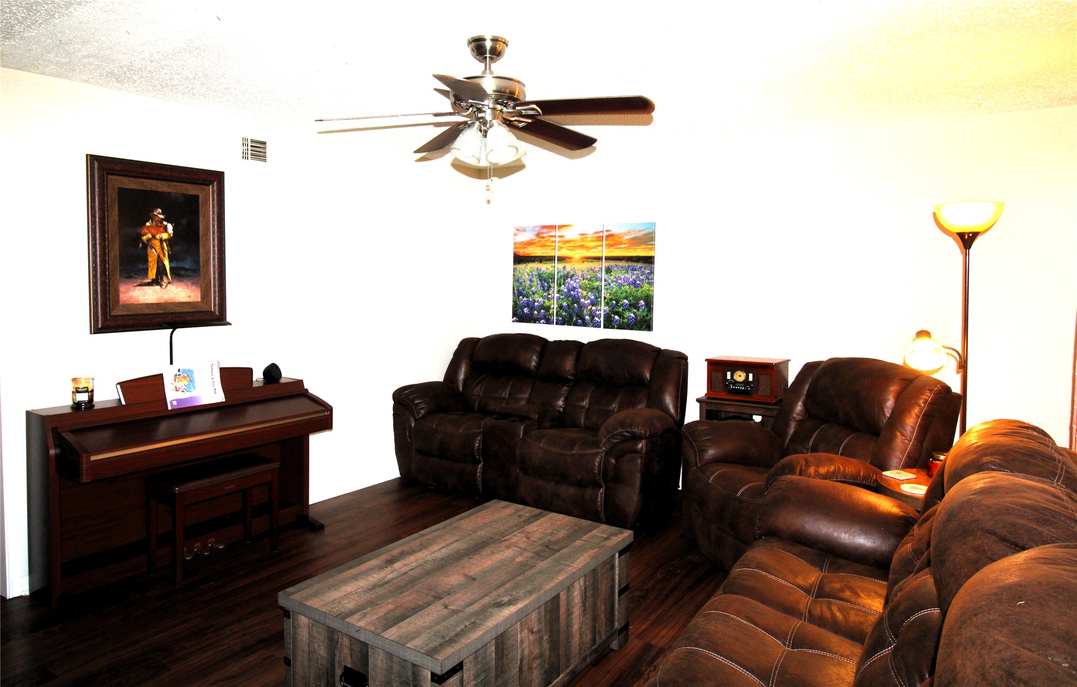 303 North 9th Street Beasley, TX 77417 - Photo 3 of 41 This cozy living room features dark leather sofas, a wooden coffee table, and a ceiling fan. It has a piano against the wall, warm lighting from floor lamps, and colorful artwork adding a welcoming touch.