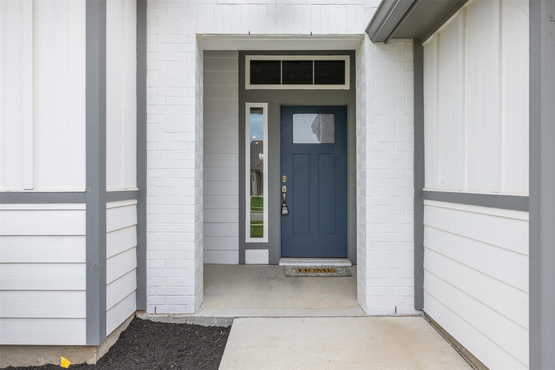 1968 Chief Street Bryan, TX 77807 - Photo 20 of 27 This photo shows a modern home entrance with a blue front door, transom window, and side window. The exterior features light-colored brick and siding with gray trim, creating a welcoming and contemporary look.
