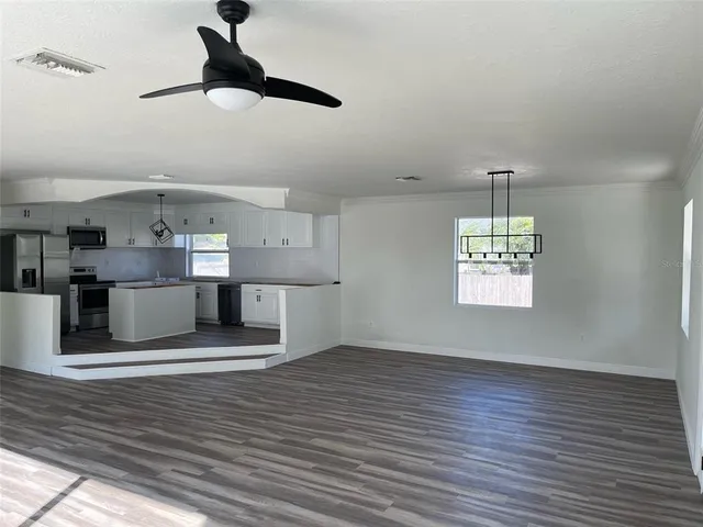 an empty room with wooden floor ceiling fan and kitchen view
