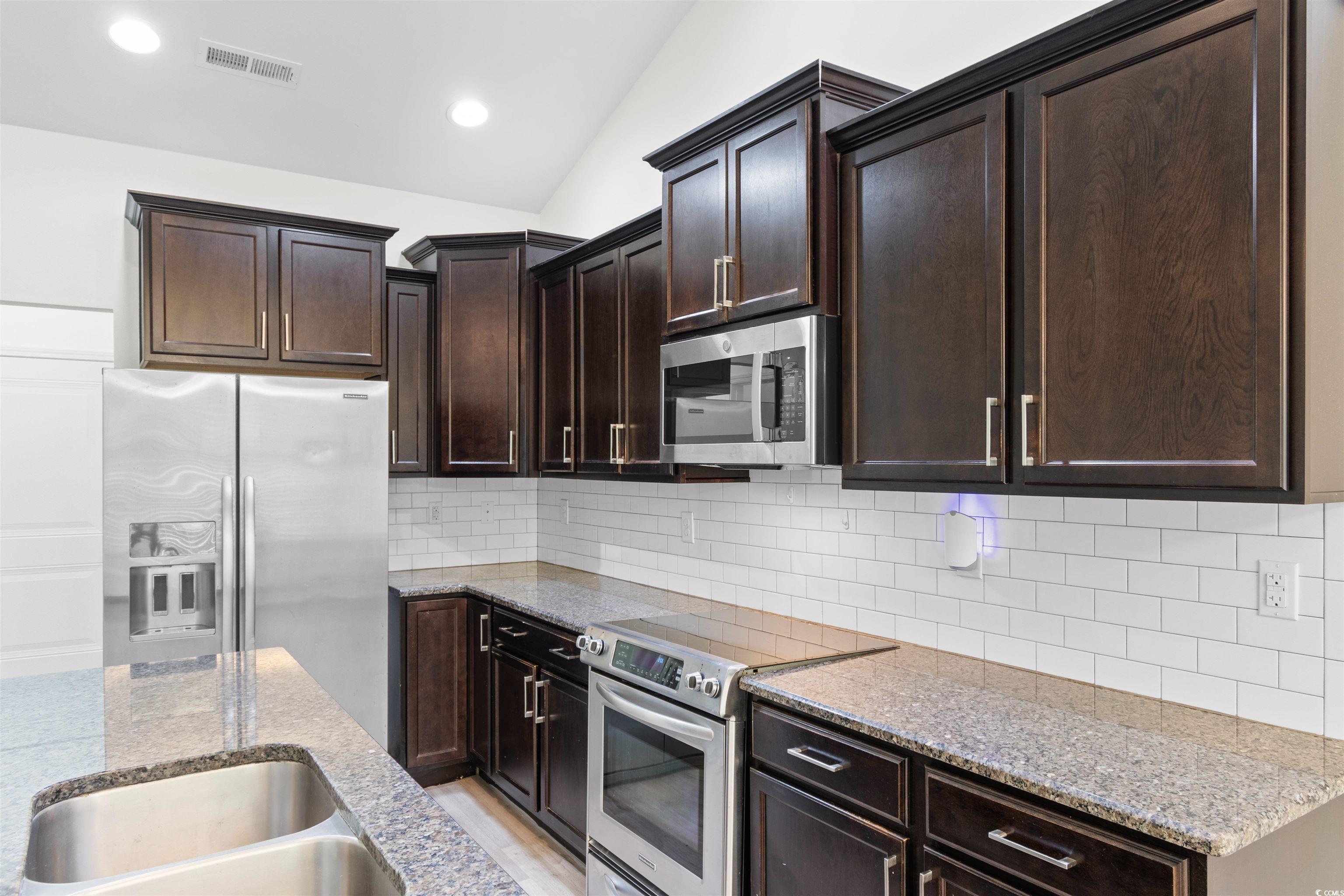 124 Shady Arbor Loop Longs, SC 29568 - Photo 5 of 40 Kitchen with appliances with stainless steel finishes, light stone countertops, dark brown cabinets, vaulted ceiling, and backsplash