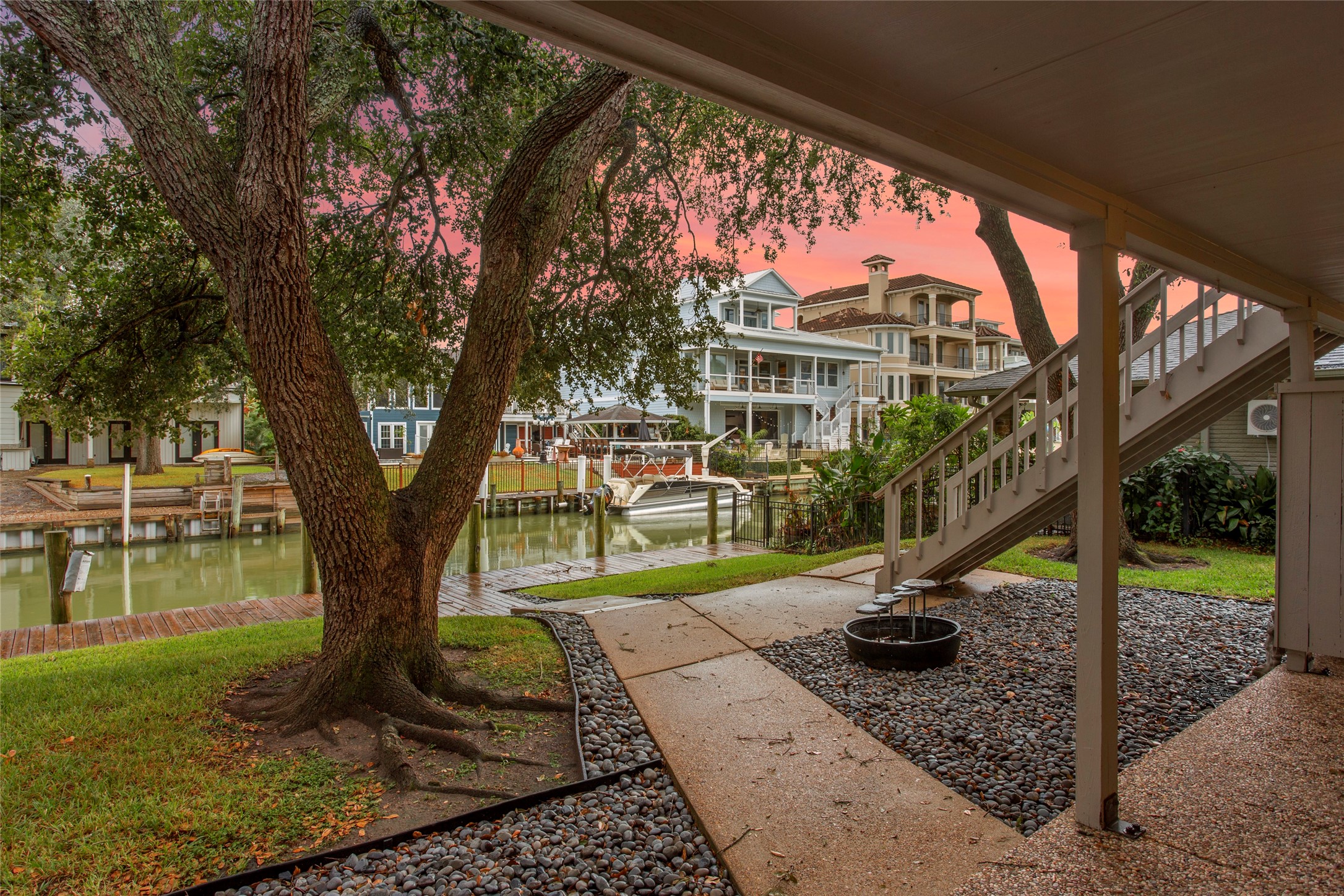 67 Harbor Lane Kemah, TX 77565 - Photo 34 of 47 This inviting waterfront property features a peaceful backyard with mature trees and water views with the sunset in the background. The cozy patio area offers a perfect spot for relaxation, with a staircase leading to the upper level. Ideal for those seeking a tranquil, scenic living environment.