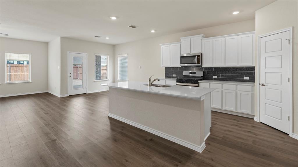 447 Epiphany Lane Lavon, TX 75166 - Photo 3 of 31 Open-concept living space featuring wood-finish flooring, a kitchen island with an integrated sink, white cabinetry, a stainless steel microwave and range, and a dark subway tile backsplash