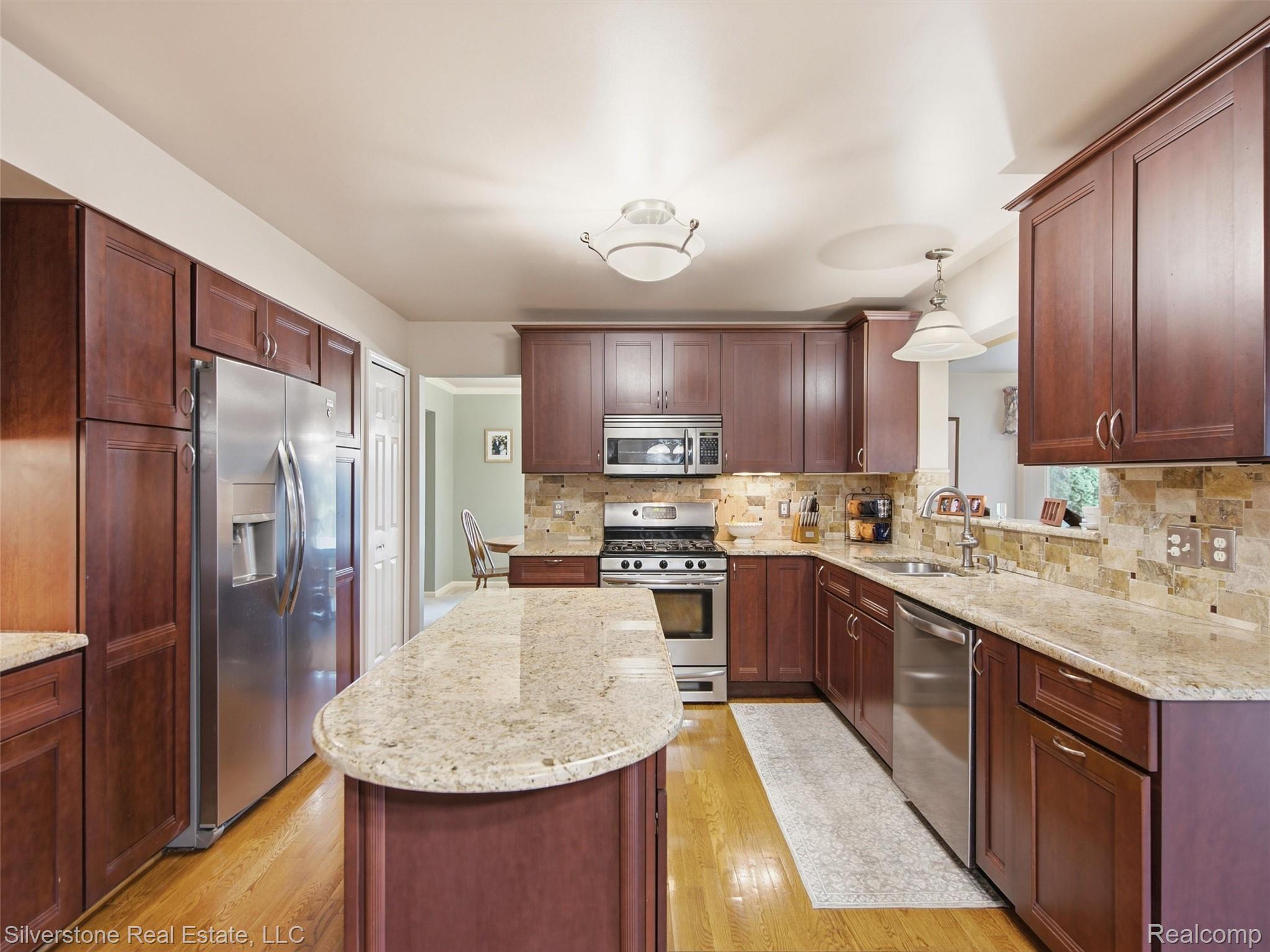 6577 Edith Court Troy, MI 48085 - Photo 22 of 61 Kitchen featuring wood-finish flooring, dark wood cabinetry, a central island with a light-toned countertop, stainless steel appliances, and a tiled backsplash