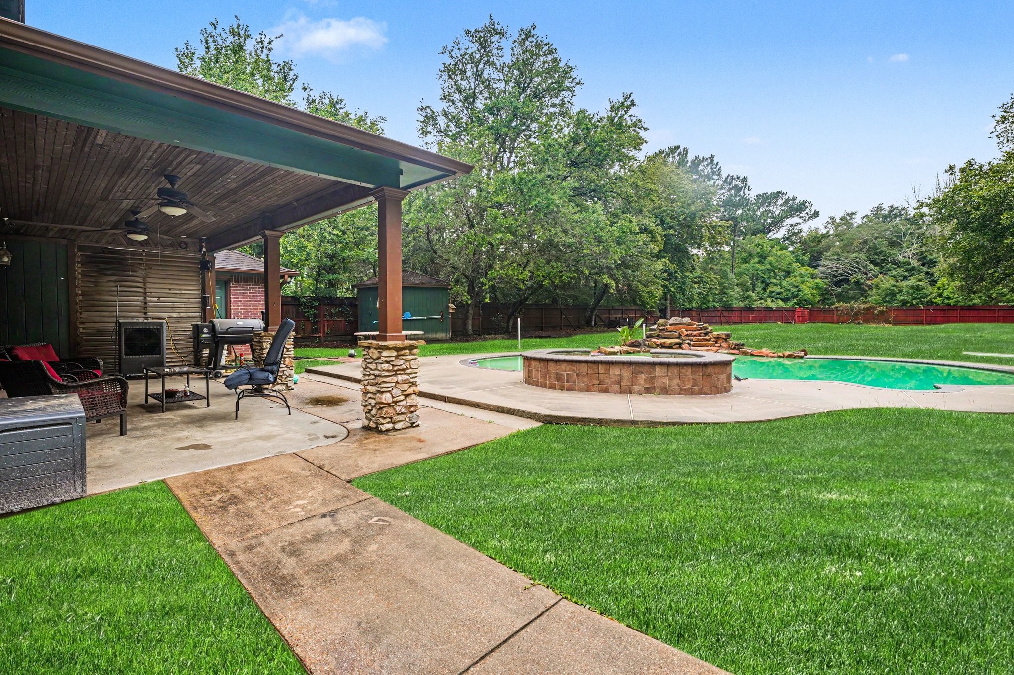 5104 Avenue R Santa Fe, TX 77510 - Photo 16 of 35 This backyard features a covered patio with ceiling fans, outdoor seating, and a fire pit. It overlooks a spacious lawn with a pool and waterfall feature, surrounded by mature trees for privacy.