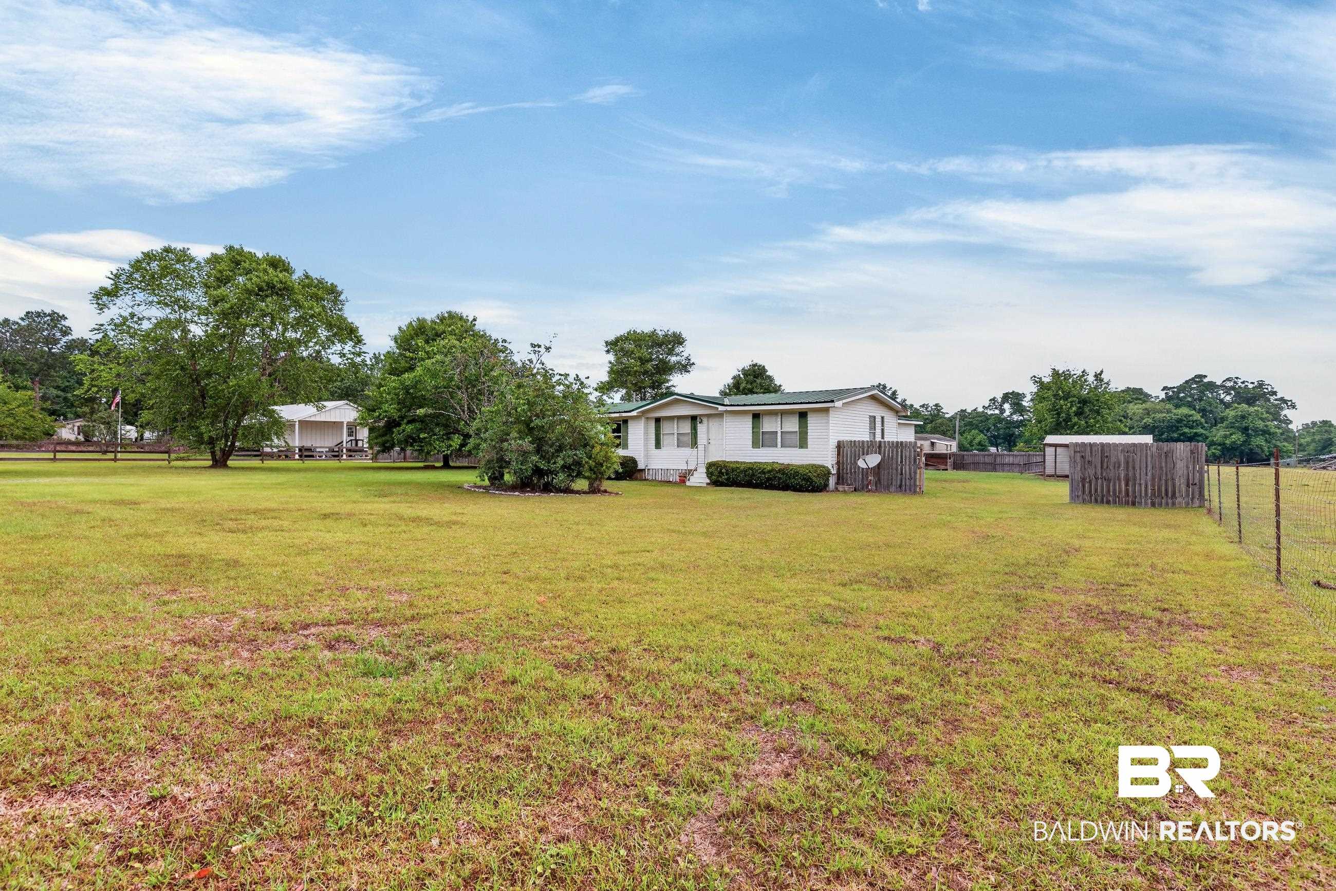 24903 Patterson Road Robertsdale, AL 36567 - Photo 6 of 57 Expansive green lawn surrounding a white residential structure with a green roof, featuring a wood fence, mature trees, and a distant outbuilding