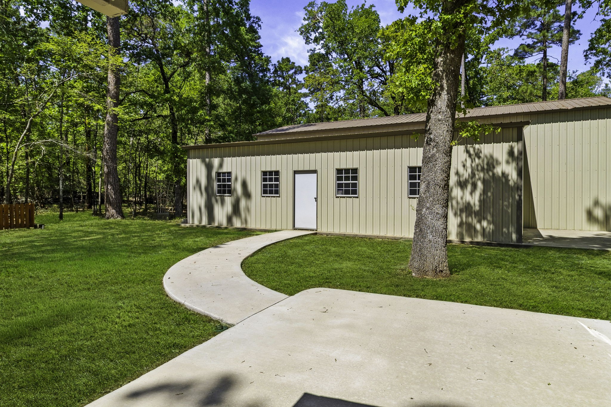 21 Red Oak Circle Point Blank, TX 77364 - Photo 27 of 32 This photo showcases a spacious metal outbuilding set in a lush, wooded backyard. It features a paved walkway and a well-maintained lawn, perfect for storage or a workshop.