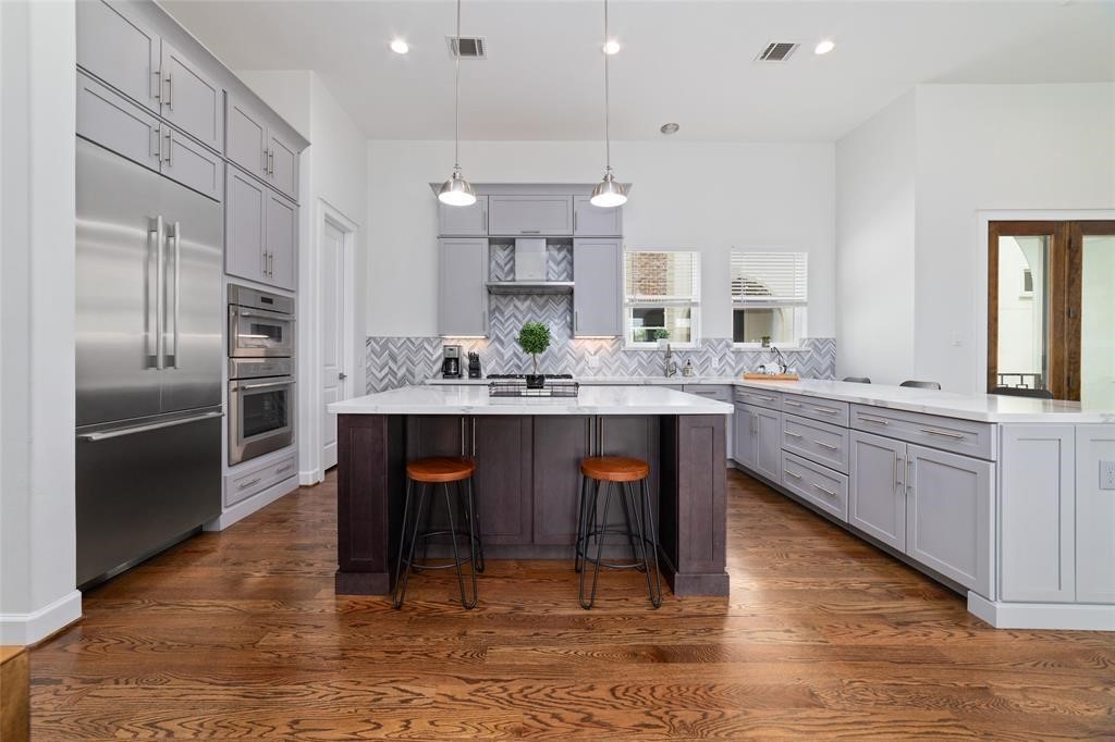 710 Delano Street, Unit A Houston, TX 77003 - Photo 5 of 32 The kitchen features modern gray cabinetry, a spacious quartz island with seating, stainless steel appliances, and hardwood floors. The backsplash has a stylish herringbone pattern, and pendant lighting adds a contemporary touch.