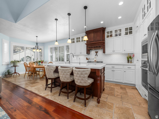 968 South Cyn Road Santaquin, UT 84655 - Photo 4 of 36 Kitchen and dining area featuring a central island with countertop seating, white cabinetry, a decorative wood range hood, stainless steel appliances, and a tiled backsplash