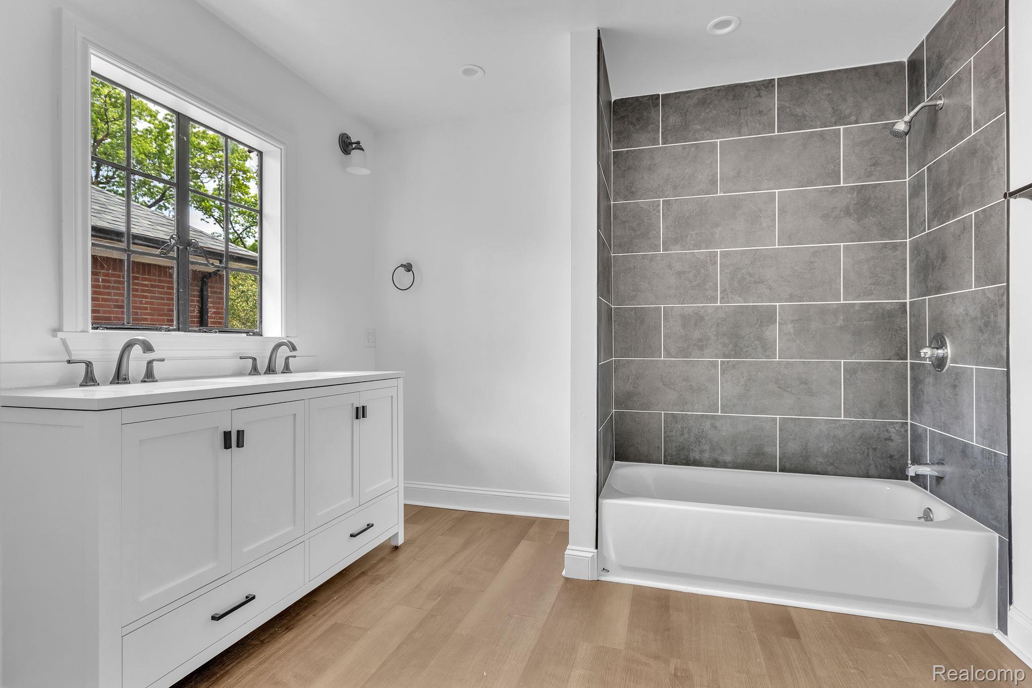 19510 Stratford Road Detroit, MI 48221 - Photo 29 of 45 Bathroom featuring a dual-sink vanity with white cabinetry and dark hardware, a bathtub with gray subway tile surround, wood-finish flooring, and a multi-pane window