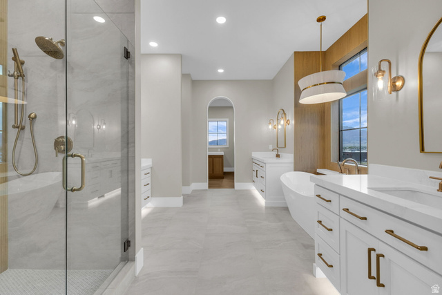 2828 Jojo Road Eagle Mountain, UT 84005 - Photo 25 of 63 Sleek bathroom featuring a glass-enclosed shower with brushed brass fixtures, a freestanding soaking tub, dual vanities with white countertops and cabinetry, and large windows framed by wood-finish accents