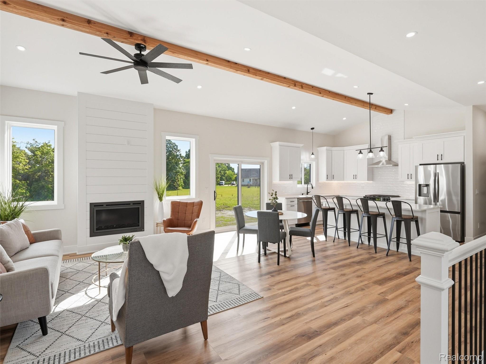 77850 Angling Road Richmond, MI 48062 - Photo 8 of 54 Vaulted ceilings with exposed wood beams, a shiplap-paneled fireplace, wood-finish flooring, white kitchen cabinetry with stainless steel appliances, and a kitchen island with bar seating
