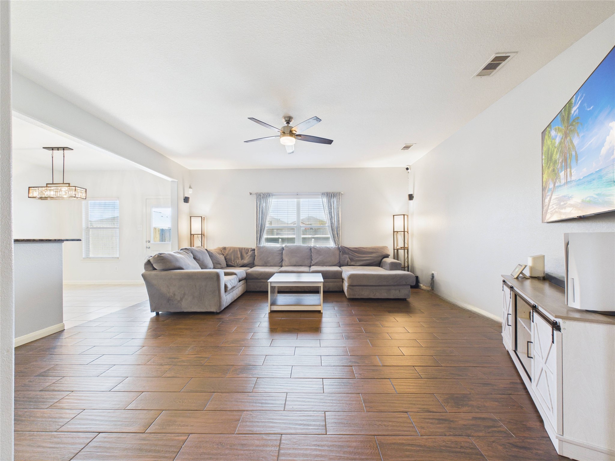 911 Marigold Road Baytown, TX 77521 - Photo 10 of 32 Bright and spacious living room with wood-look tile flooring, a large sectional sofa, ceiling fan, and a wall-mounted TV. It opens to a dining area with natural light and modern fixtures, creating an inviting atmosphere.