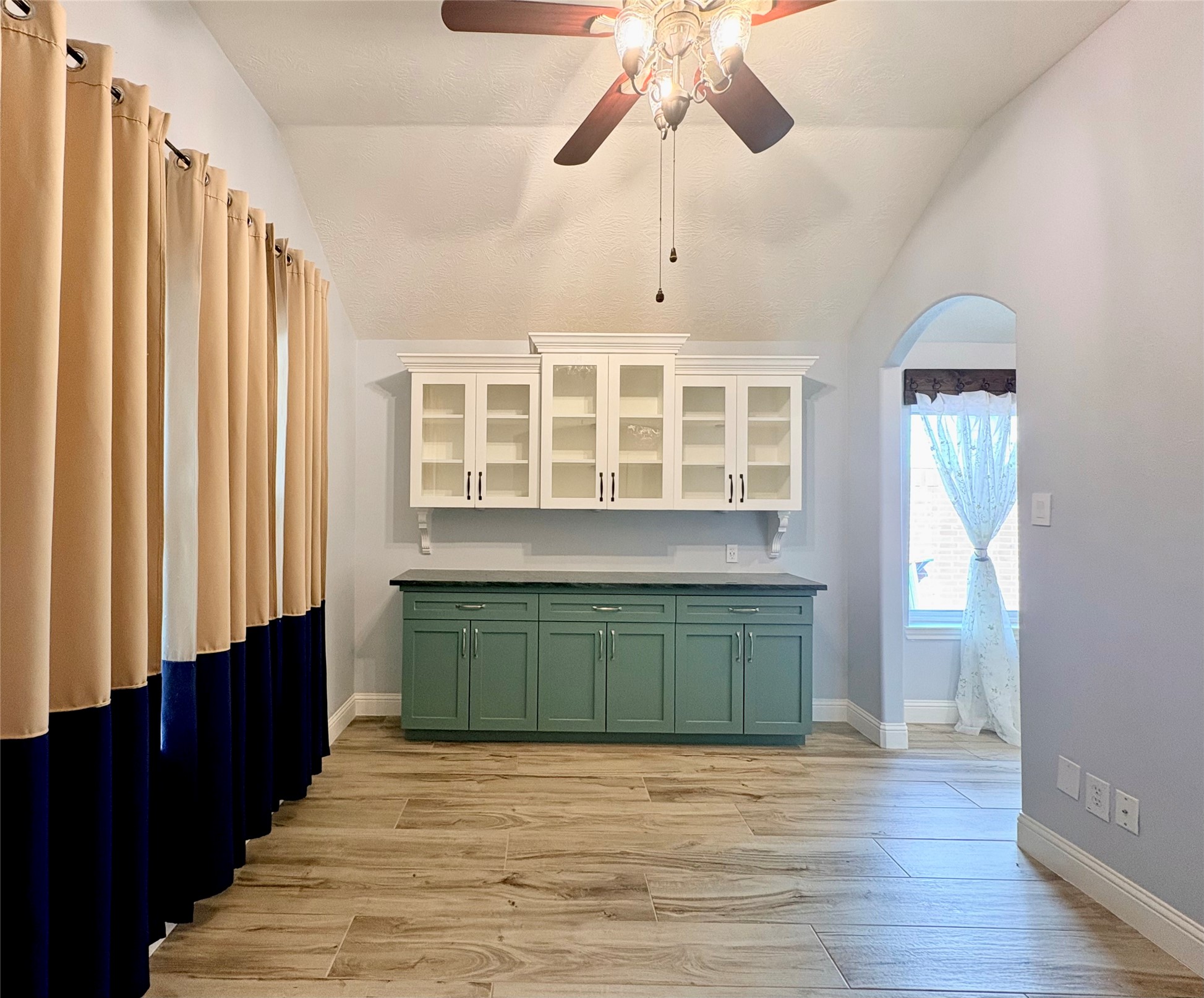 15410 Redbud Berry Way Cypress, TX 77433 - Photo 6 of 49 This room features high ceilings with a ceiling fan, a built-in cabinet with glass doors, and a mix of green and white cabinetry. It has large windows with curtains, letting in natural light, and wood-patterned flooring. Perfect for a cozy living space or study.