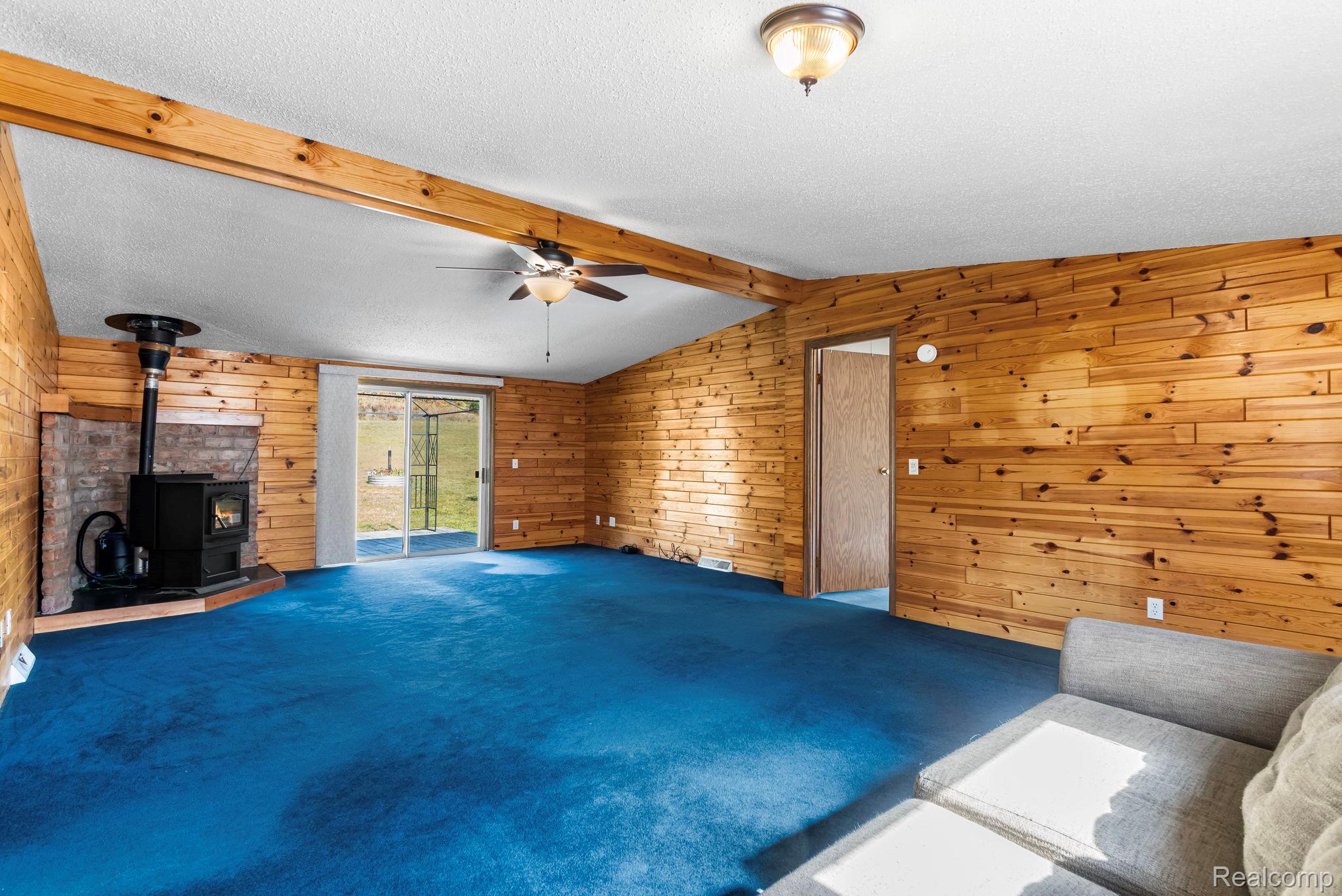 Wood-paneled interior walls, exposed wood beams, a wood-burning stove with a stone surround, and a ceiling fan