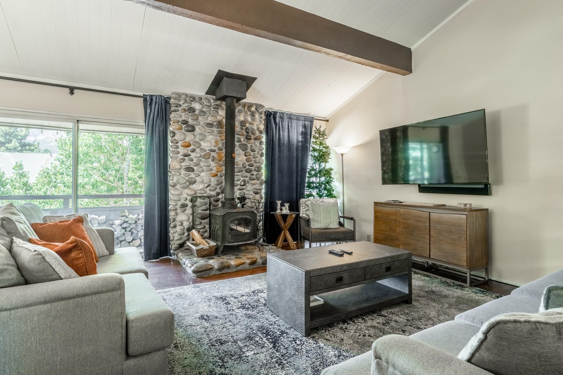 Living area featuring a floor-to-ceiling river stone fireplace with a wood-burning stove, vaulted ceilings with exposed wood beams, and wood-finish flooring