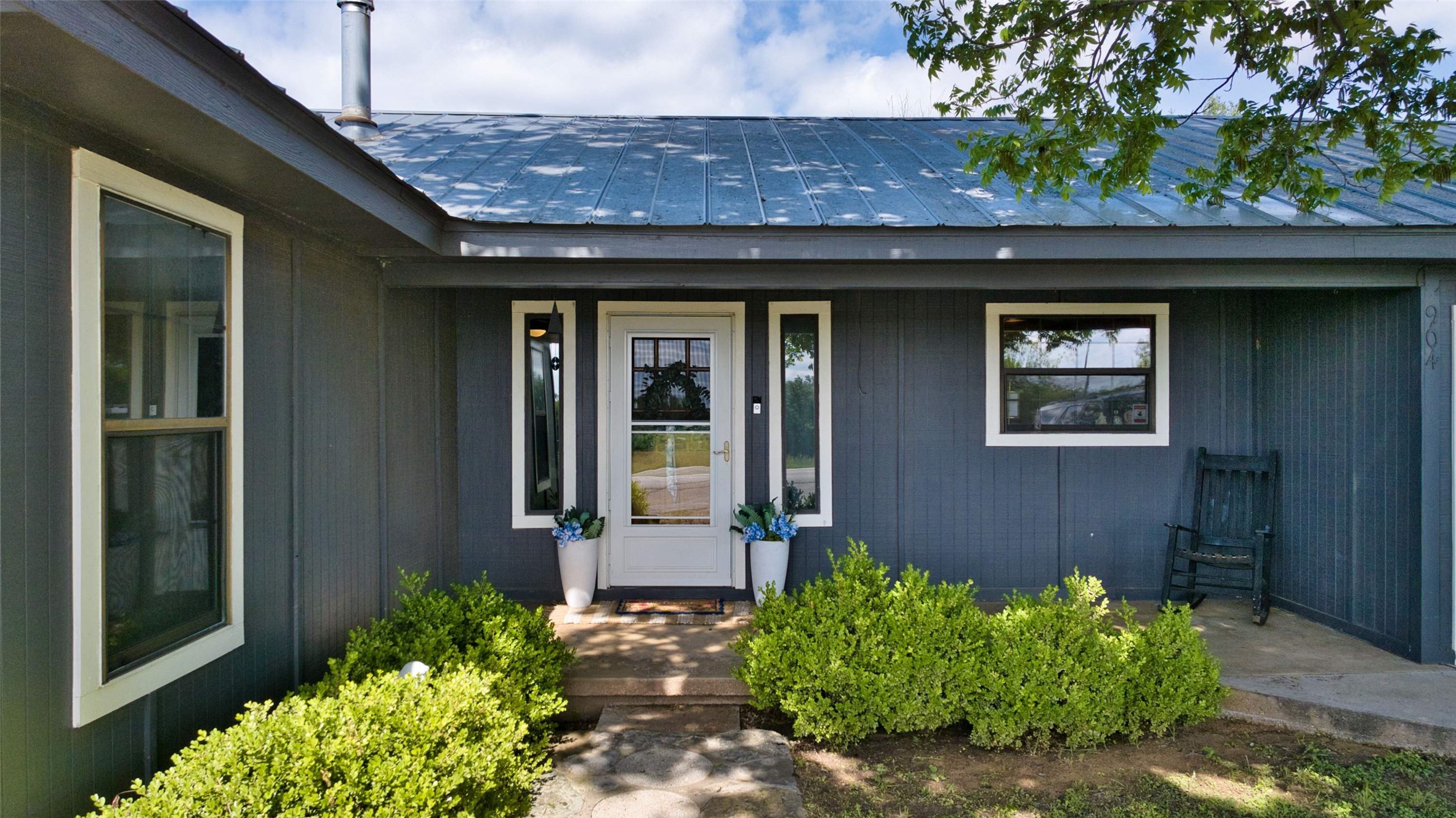 904 Bluebonnet Road Mason, TX 76856 - Photo 2 of 28 Exterior featuring a dark blue vertical siding, a metal roof, white trim windows, a white entry door with glass panels, and established shrubbery