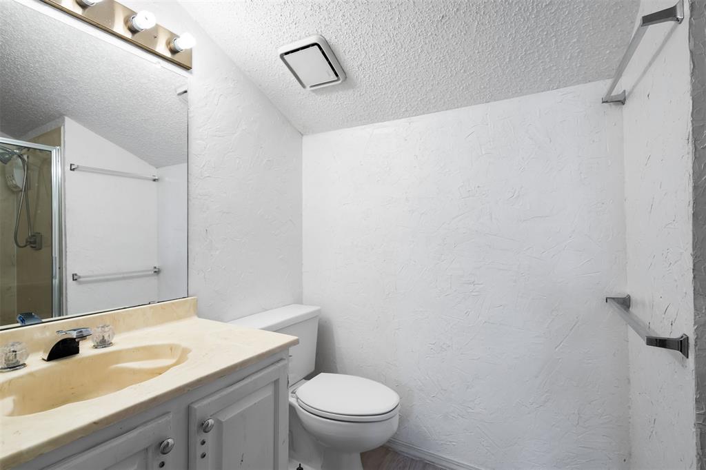 5590 Spring Valley Road, Unit H 203 Dallas, TX 75254 - Photo 21 of 27 Bathroom featuring a vanity with a beige countertop and integrated sink, a built-in toilet, a glass-enclosed shower with a chrome showerhead, and wall-mounted towel bars
