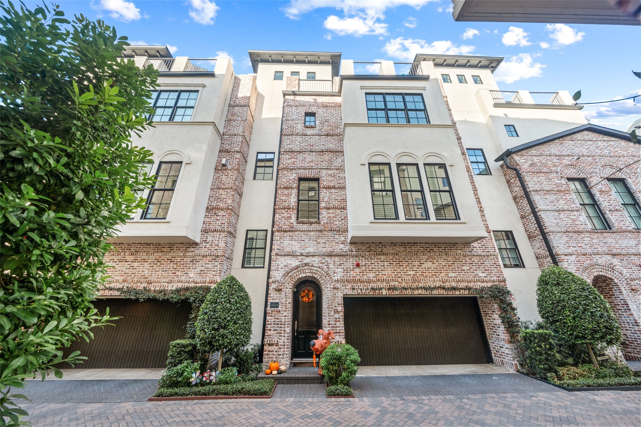 Modern townhouse with a blend of brick and stucco exterior. It features large windows, a two-car garage, and well-maintained landscaping with topiary shrubs. The design is elegant and contemporary, suitable for comfortable urban living.