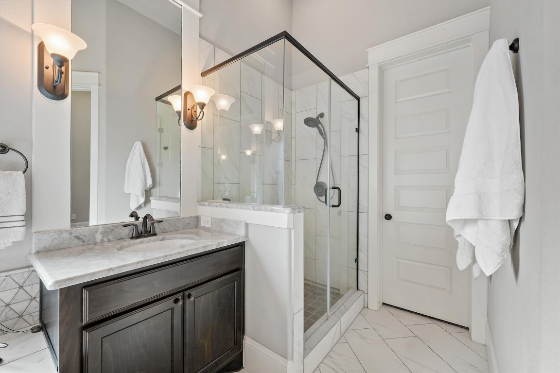 2172 Rope Maker Road Conroe, TX 77384 - Photo 23 of 50 This modern bathroom features a sleek glass-enclosed shower with a stylish black frame and fixtures. It has a marble countertop on a dark wood vanity, complemented by elegant wall sconces. The light-colored walls and floor tiles create a bright and spacious spa feel.