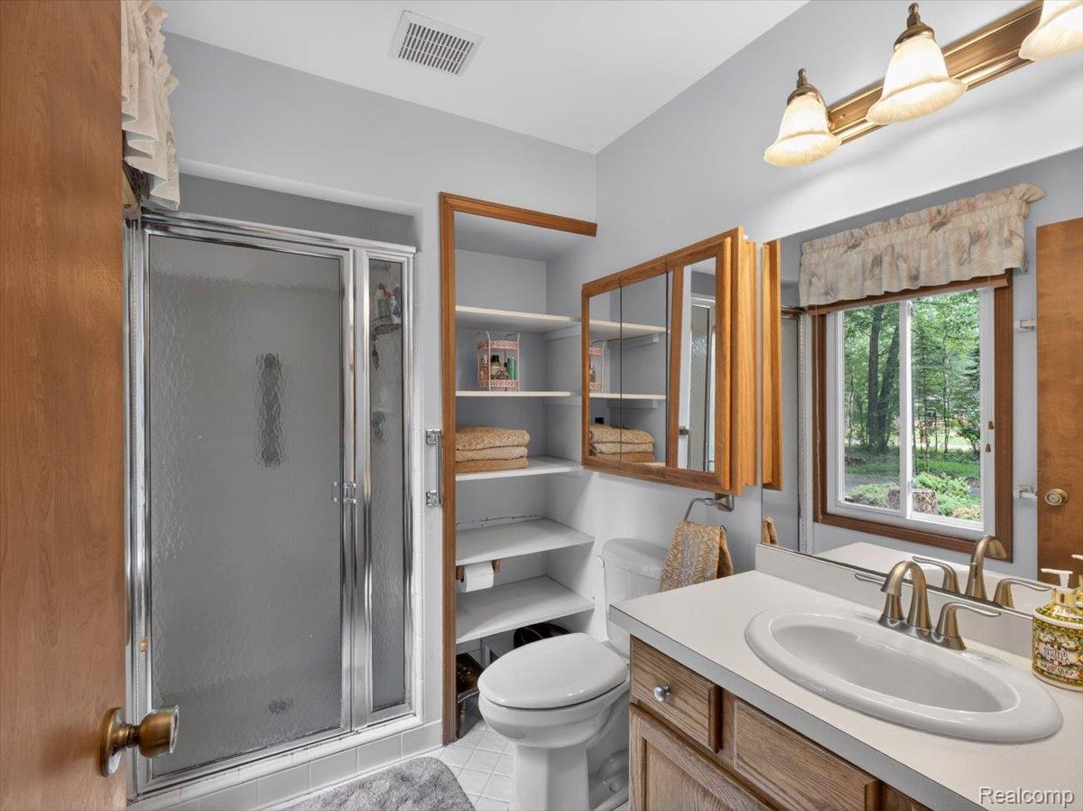 9714 Naiad Road Clarkston, MI 48348 - Photo 34 of 61 Primary ensuite bathroom offers a glass-enclosed step-in ceramic tile shower, a vanity with a white countertop and integrated sink, a wood-framed medicine cabinet, and thoughtfully designed built-in shelving for added storage.