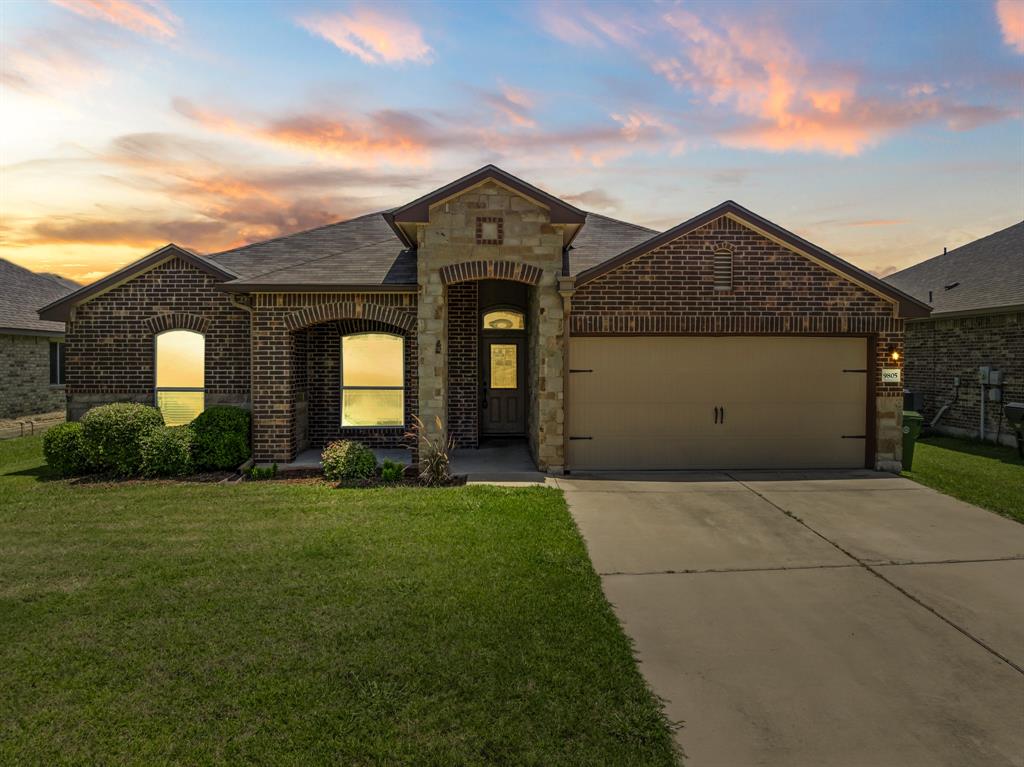 French country inspired facade featuring driveway, brick siding, an attached garage, and a front lawn