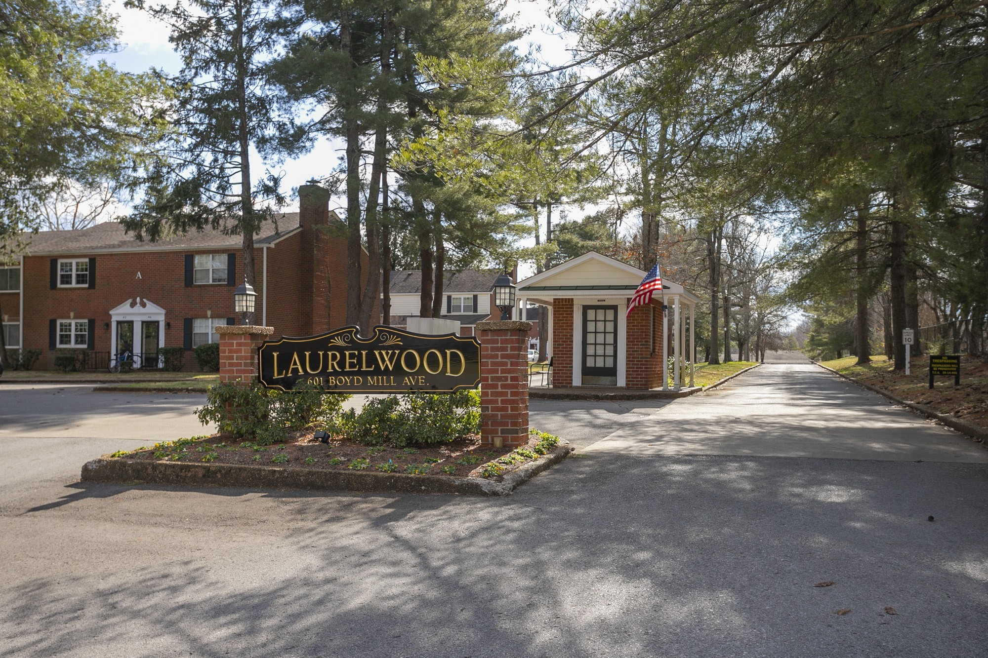a sign board with large trees and cars parked on road