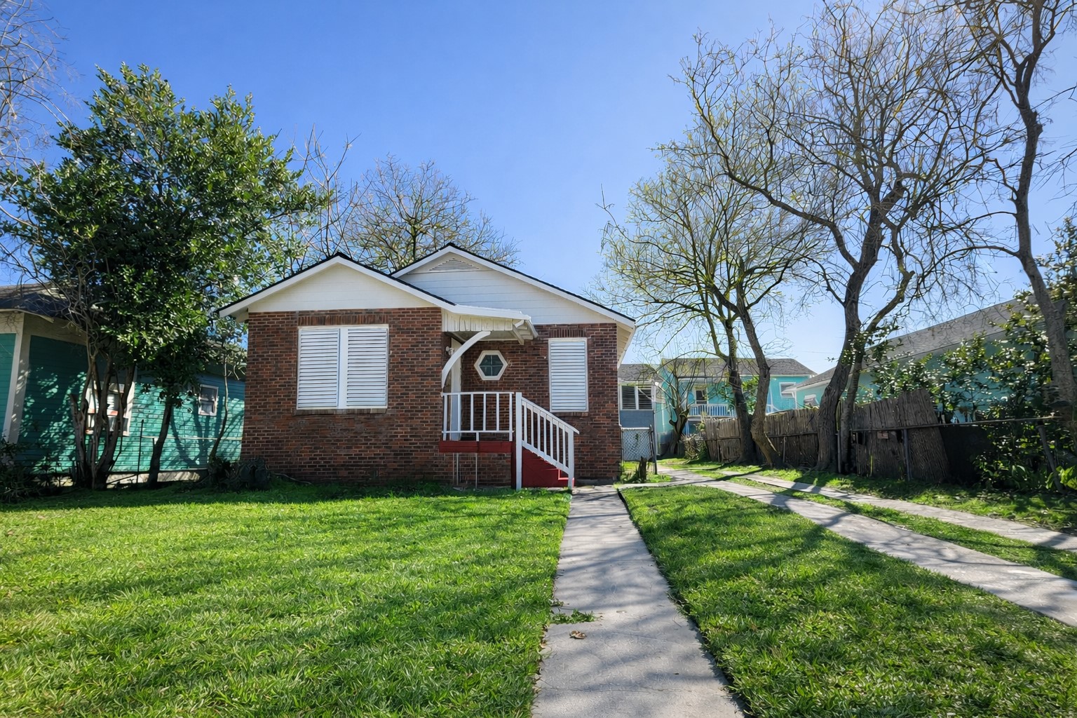 4917 Avenue R 1/2 Galveston, TX 77551 - Photo 1 of 21 Centered front view of single-story brick home with white trim, red steps, concrete walkway, green lawn, and mature trees.
