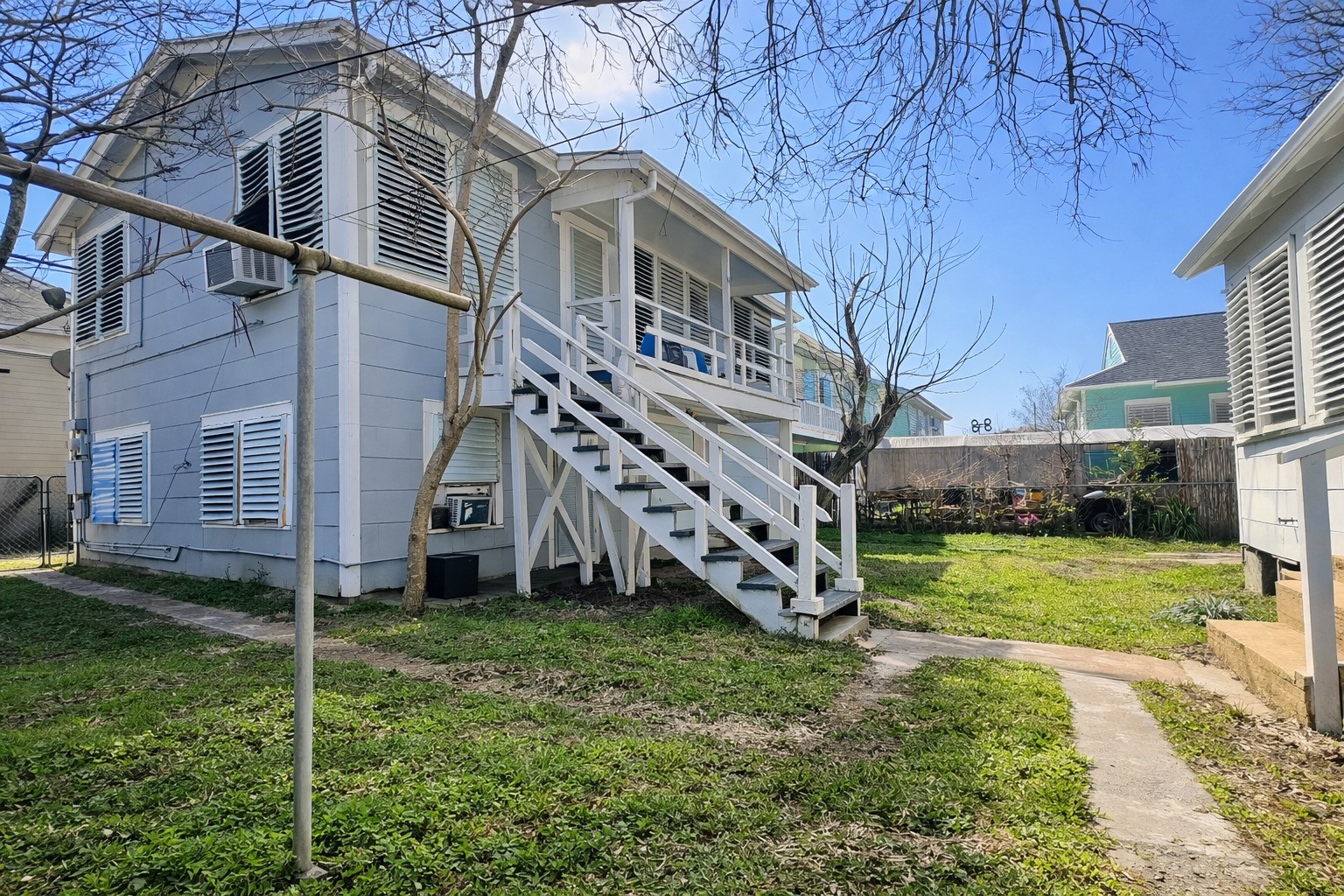 4917 Avenue R 1/2 Galveston, TX 77551 - Photo 20 of 21 Backyard view of raised blue home with exterior staircase, white railings, shuttered windows, and grassy yard between neighboring homes.