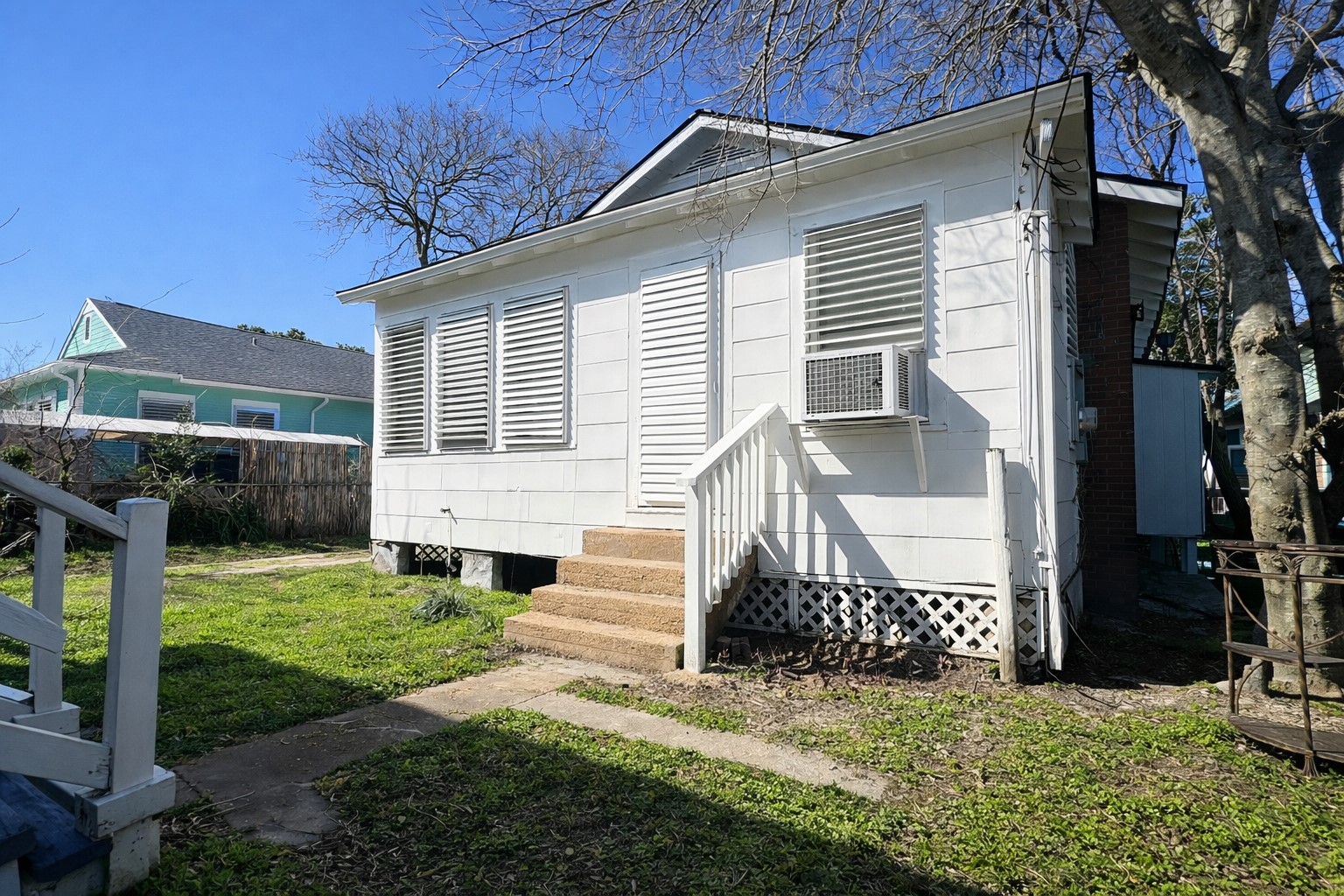 4917 Avenue R 1/2 Galveston, TX 77551 - Photo 5 of 21 Side view of white cottage-style home with louvered shutters, small staircase, window AC unit, and grassy yard under clear blue sky.