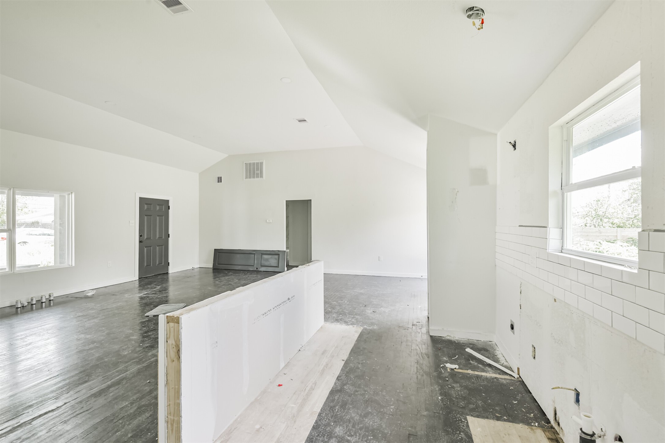 209 Leago Street Houston, TX 77022 - Photo 9 of 24 This open-concept living space features high ceilings, large windows for natural light, and a partially finished kitchen area with white subway tile backsplash. The dark flooring contrasts with the light walls, creating a modern aesthetic.