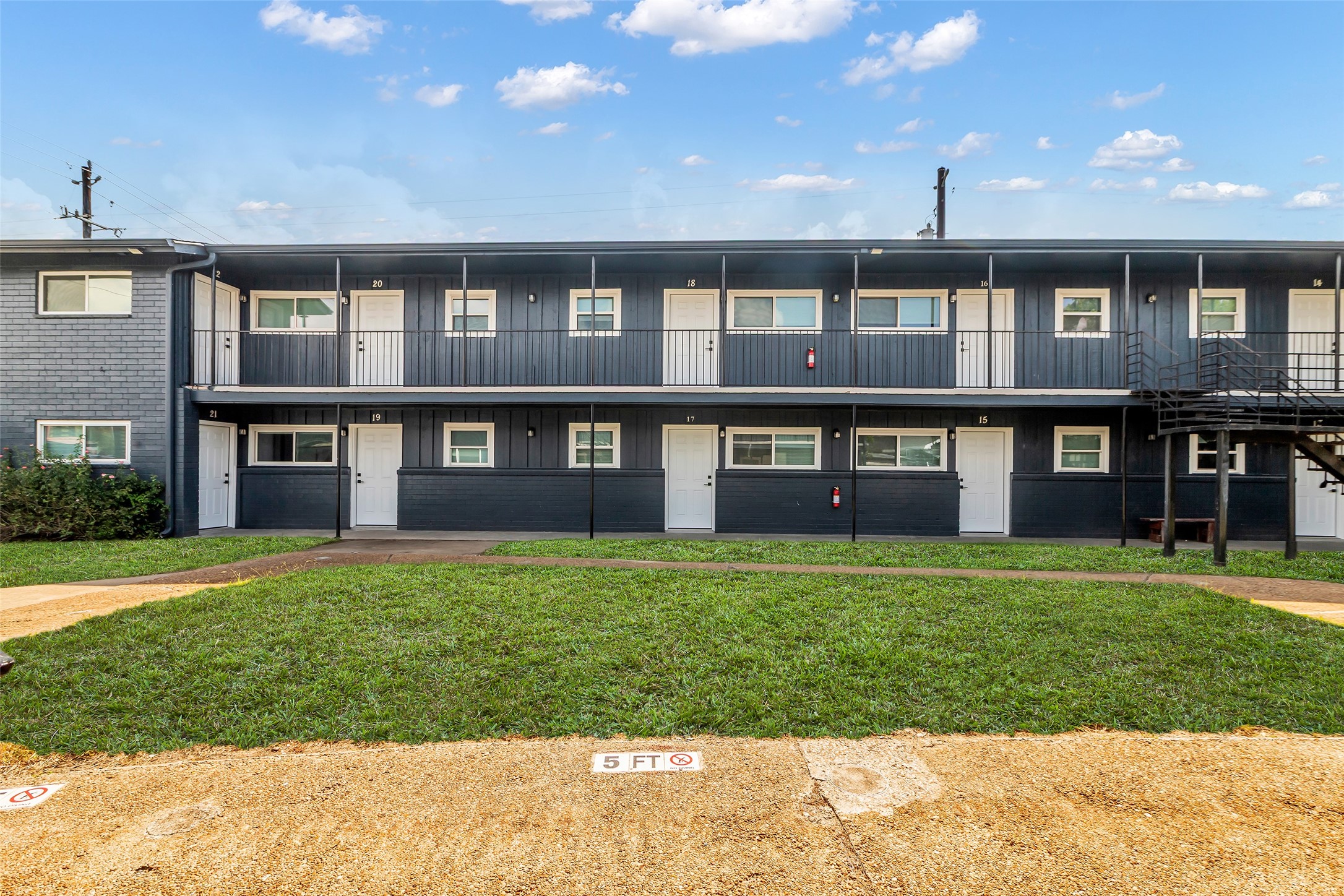 4706 Yellowstone Boulevard, Unit 21 Houston, TX 77021 - Photo 2 of 24 This photo showcases a two-story apartment building with a dark gray exterior, featuring multiple units with white doors and windows. There's a small grassy area in front, and an external staircase provides access to the upper level.