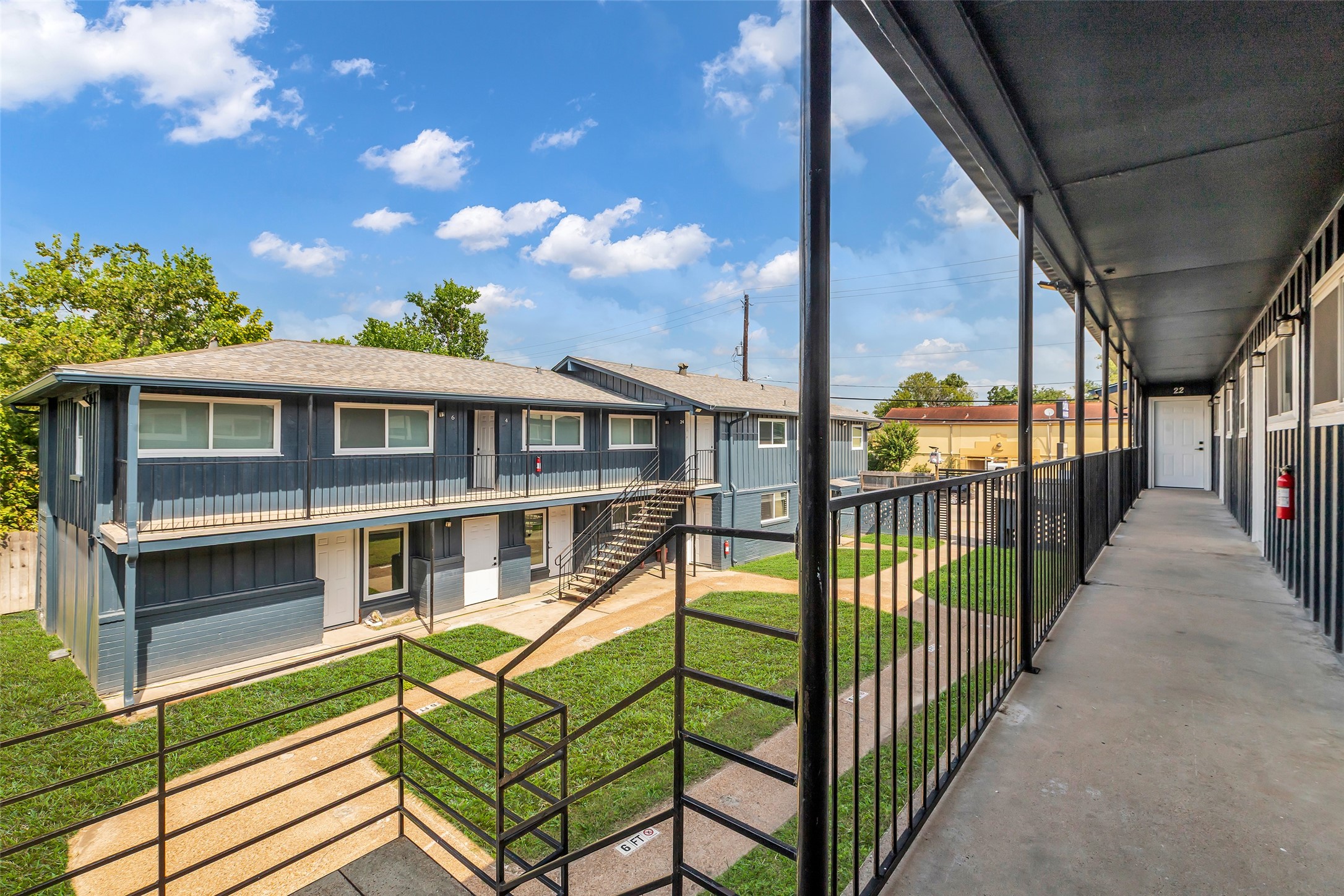 4706 Yellowstone Boulevard, Unit 21 Houston, TX 77021 - Photo 7 of 24 This photo shows a two-story apartment complex with a blue exterior and a row of windows on both levels. It features a shared walkway on the upper floor, accessible by stairs, and overlooks a well-maintained grassy courtyard. The setting is bright with clear blue skies.