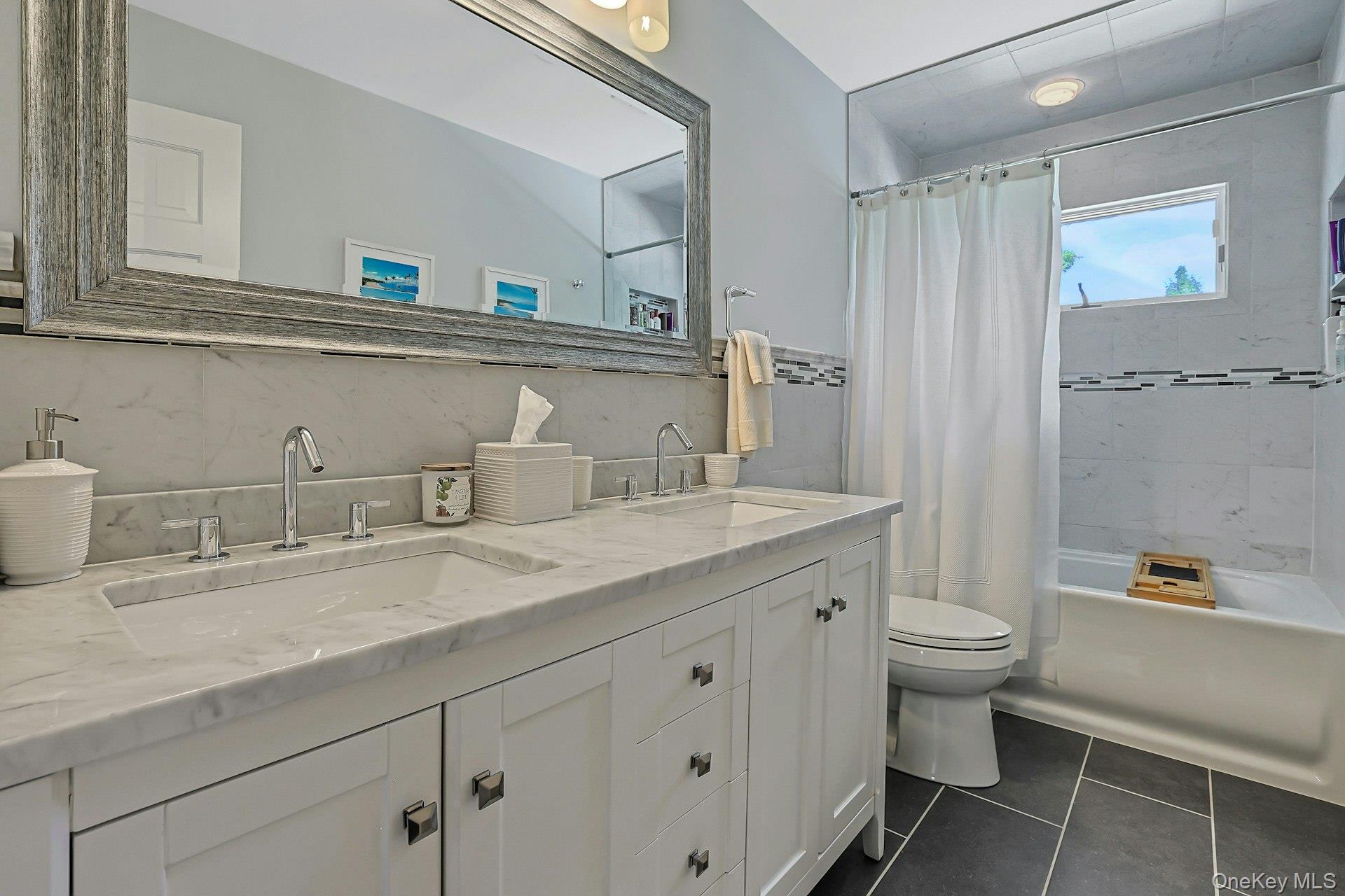 9 Stonegate Road Valhalla, NY 10595 - Photo 23 of 42 Dual vanity with white cabinetry and chrome hardware, white stone countertop, white tiled bath surround with decorative accent tile, and dark tile flooring