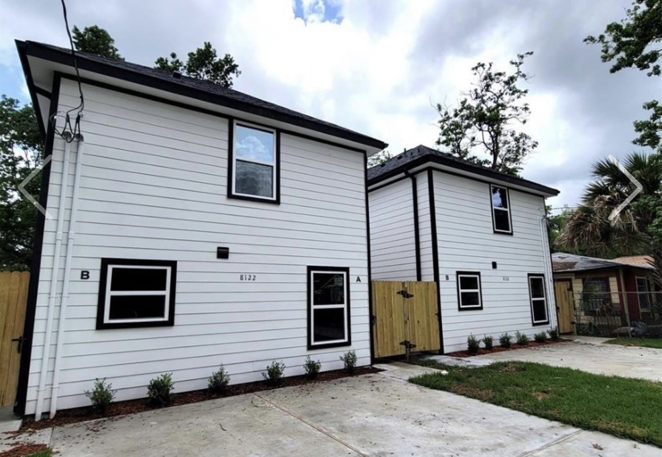 Modern duplex with a clean, white exterior and black trim. Each unit has a separate entrance, a small front yard with new landscaping, and a shared driveway. The property is surrounded by greenery, providing a fresh and inviting atmosphere.