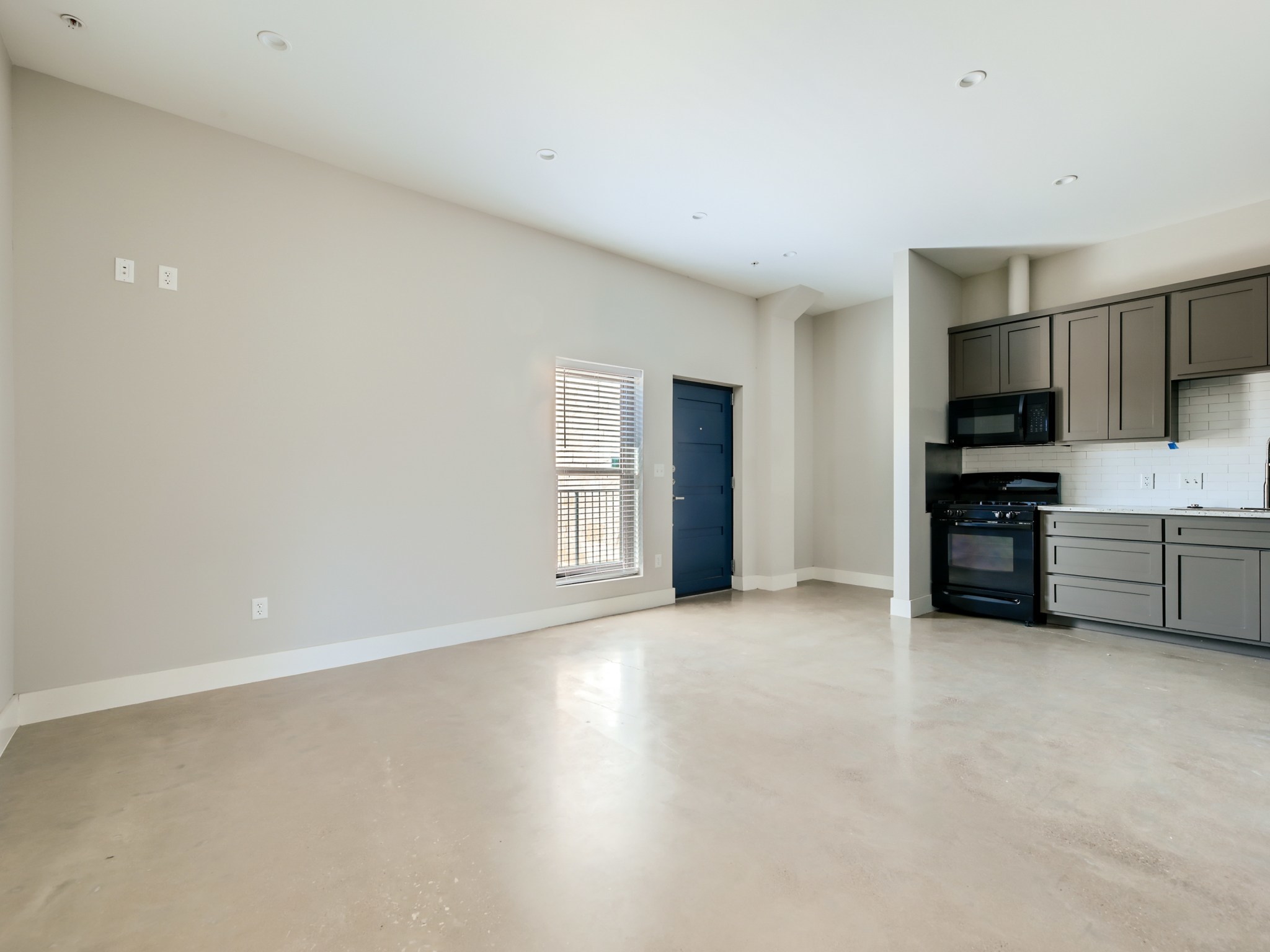 3523 Gonzalez Street, Unit 200 Austin, TX 78702 - Photo 3 of 10 Open-concept living space featuring sleek concrete flooring, recessed lighting, a modern kitchen with gray cabinetry, black appliances, and white subway tile backsplash, and a blue entry door with a window offering natural light