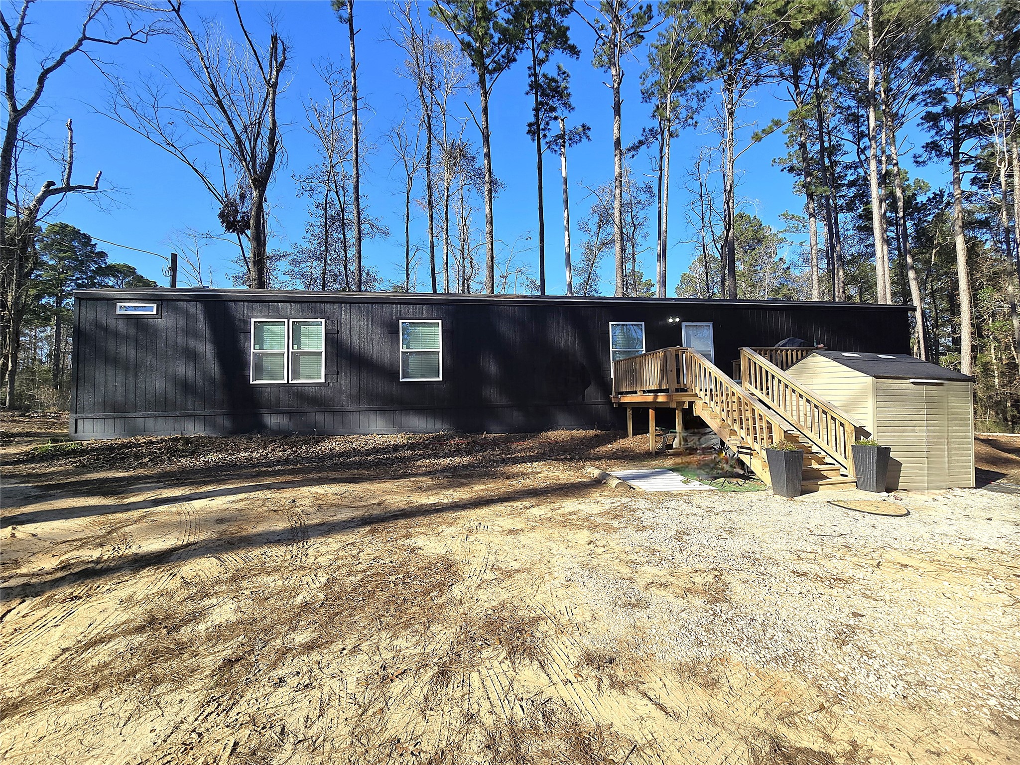 This photo shows a modern mobile home with a dark exterior, set in a wooded area. It features a small porch with stairs leading up to the entrance and a storage shed nearby, offering a serene and private setting.