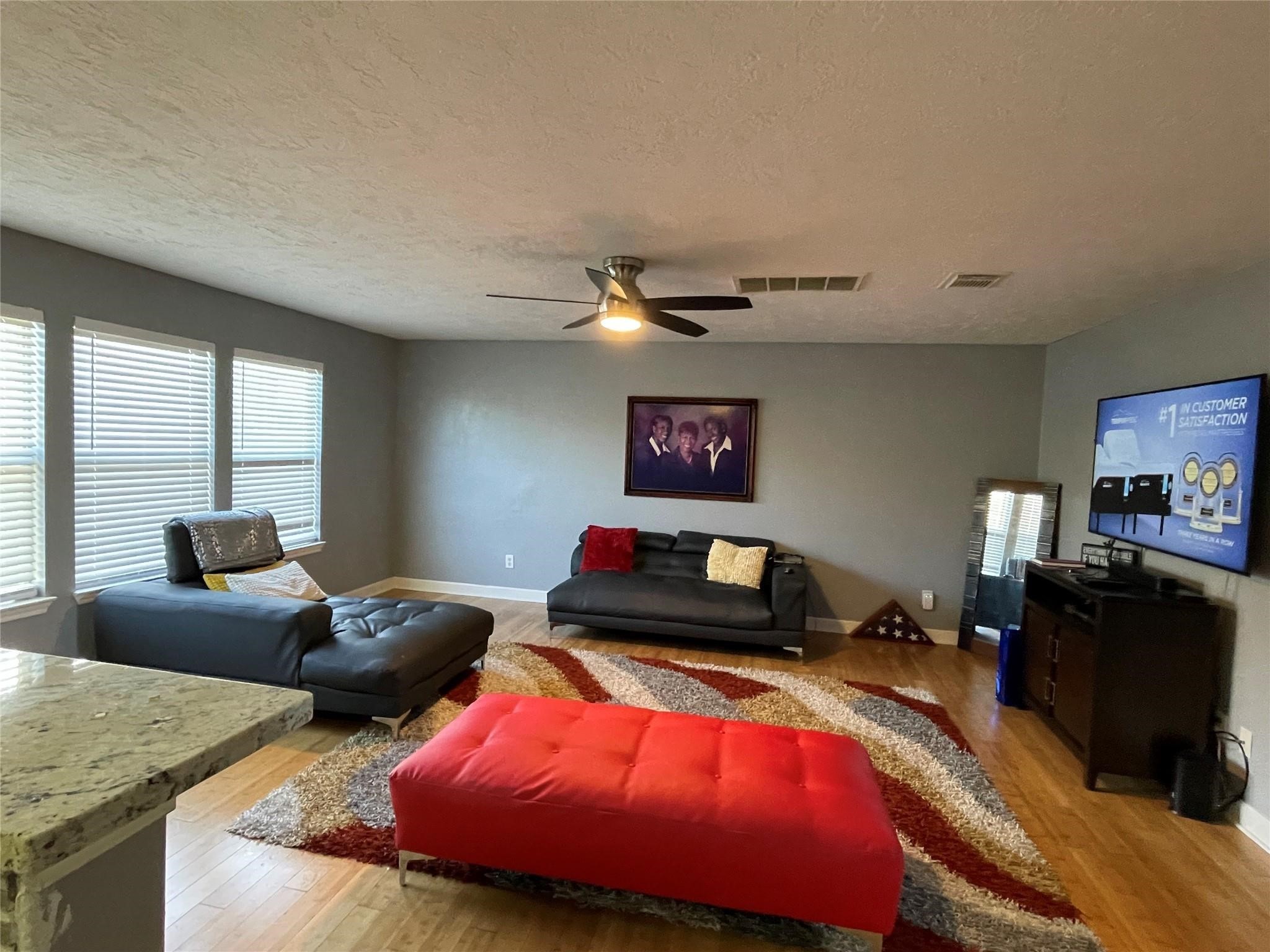 9418 Fillmont Lane Houston, TX 77044 - Photo 2 of 11 Spacious living room with hardwood floors, modern ceiling fan, and ample natural light from multiple windows. Features a sleek sectional sofa, vibrant area rug, and wall-mounted TV, creating a cozy and inviting atmosphere.