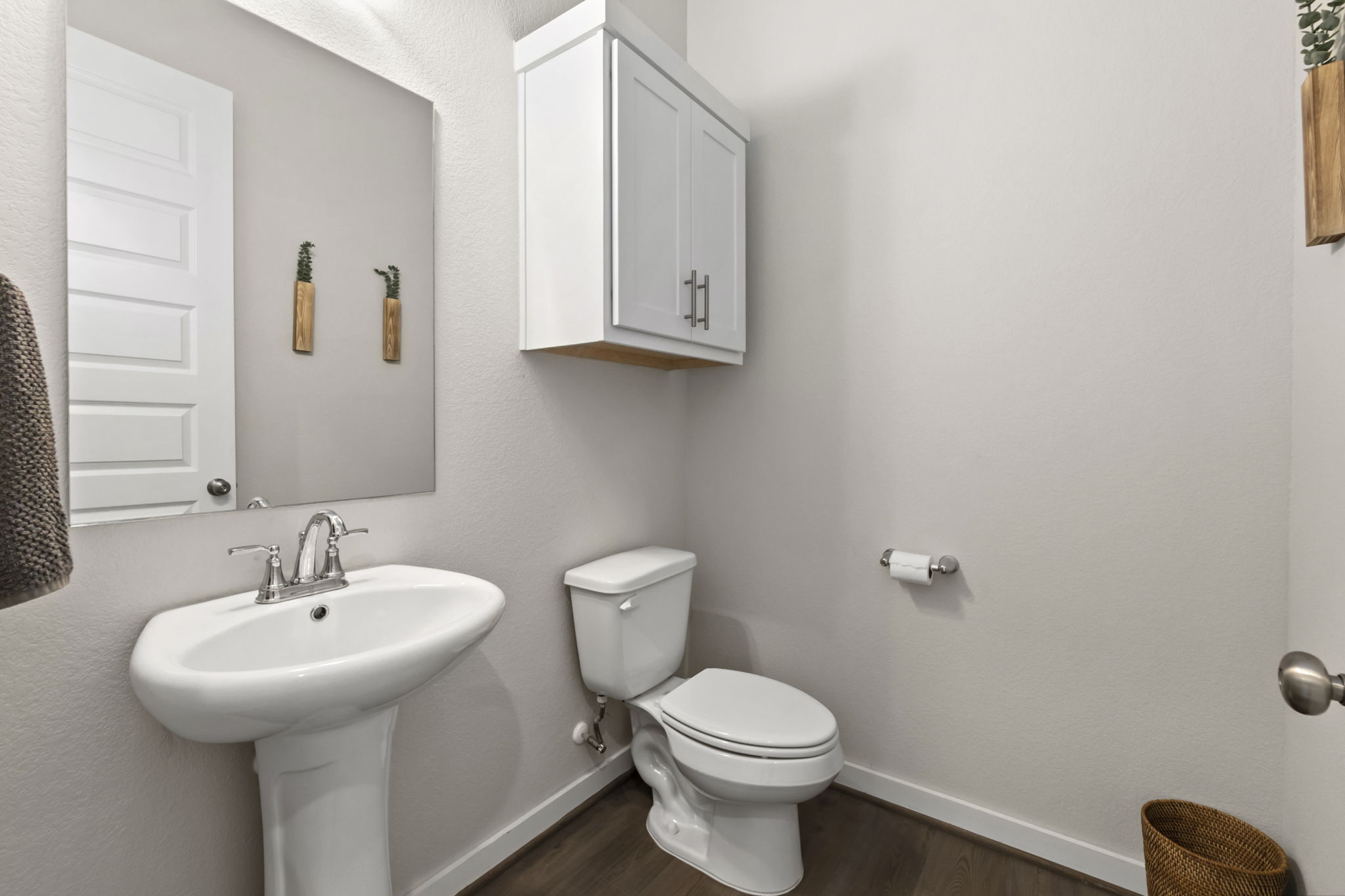 460 Constitution Way Kyle, TX 78640 - Photo 17 of 40 Pedestal sink with chrome faucet, a rectangular wall-mounted mirror, white paneled door, wood-finish flooring, and a wall-mounted white cabinet with chrome hardware