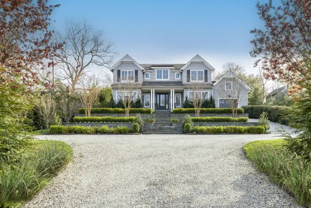 a view of a white house in front of a big yard with plants and large trees