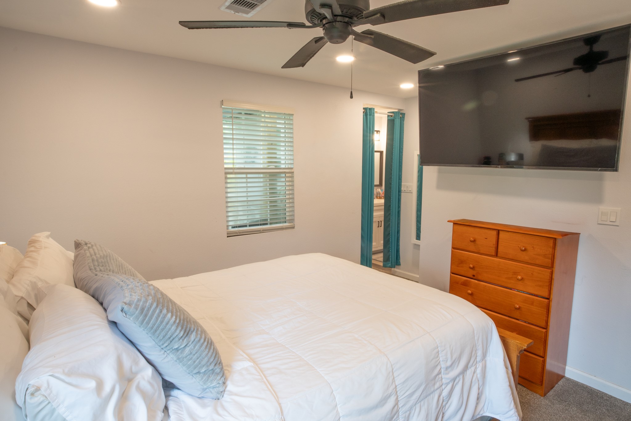 846 South Anderson Street Angleton, TX 77515 - Photo 14 of 30 This cozy bedroom features a large bed with a ceiling fan, and a mounted TV. There's a wooden dresser for storage and an ensuite bathroom visible through an open doorway. Soft lighting and a window with blinds add a warm touch.