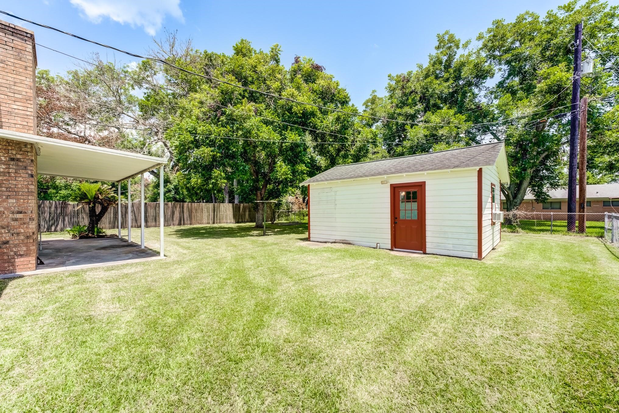 305 Yaupon Street Sweeny, TX 77480 - Photo 11 of 22 This photo showcases a spacious backyard with well-maintained grass, featuring a small outbuilding with a red door and white siding. There's a covered patio attached to a brick structure, and mature trees provide shade and privacy.