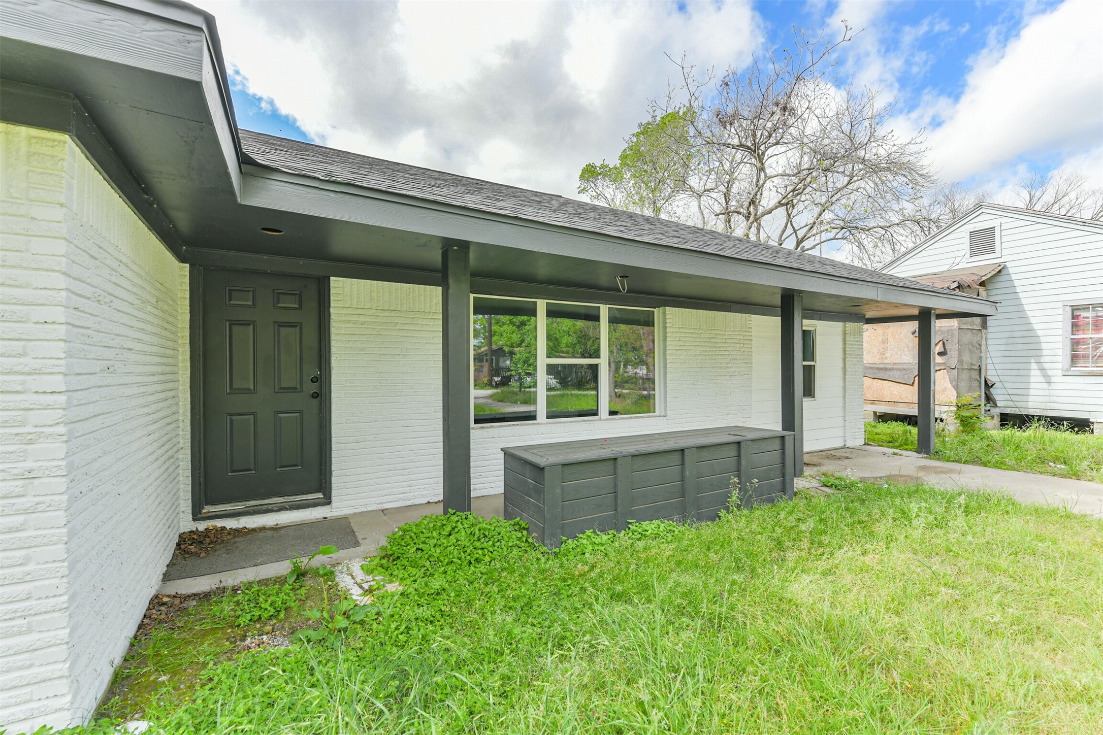 209 Leago Street Houston, TX 77022 - Photo 24 of 24 This photo showcases a single-story home with a covered front porch. The exterior features white brick with dark trim, and the entrance has a solid dark door. The surrounding lawn is grassy with a mix of greenery.