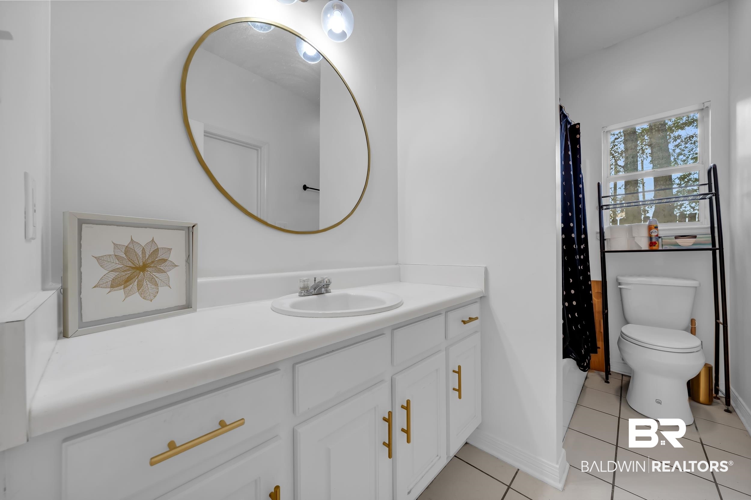 17488 Pandion Ridge, Unit A1 Fairhope, AL 36532 - Photo 16 of 18 Bathroom featuring a white vanity with gold-tone hardware, a circular mirror, and a wall-mounted light fixture