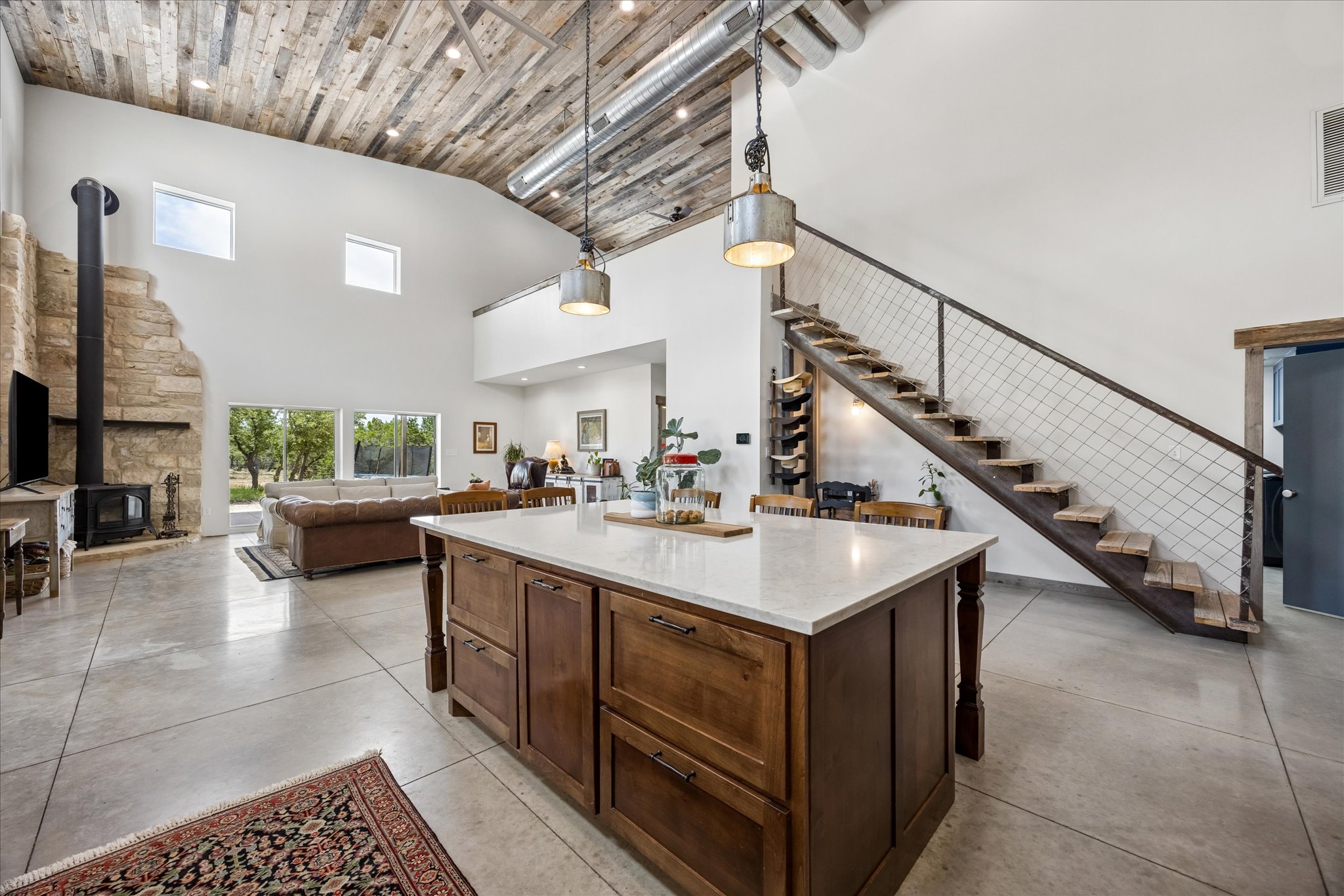6411 West Fitzhugh Road Dripping Springs, TX 78620 - Photo 9 of 34 Expansive open-concept living area featuring a large kitchen island with a vaulted wood-paneled ceiling, a stone fireplace with a wood-burning stove, concrete flooring, and an open-tread staircase with cable railing