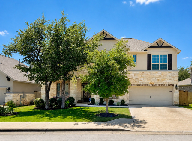 A well-maintained facade features light-colored stone and stucco, framed by mature shade trees and a vibrant green lawn.
